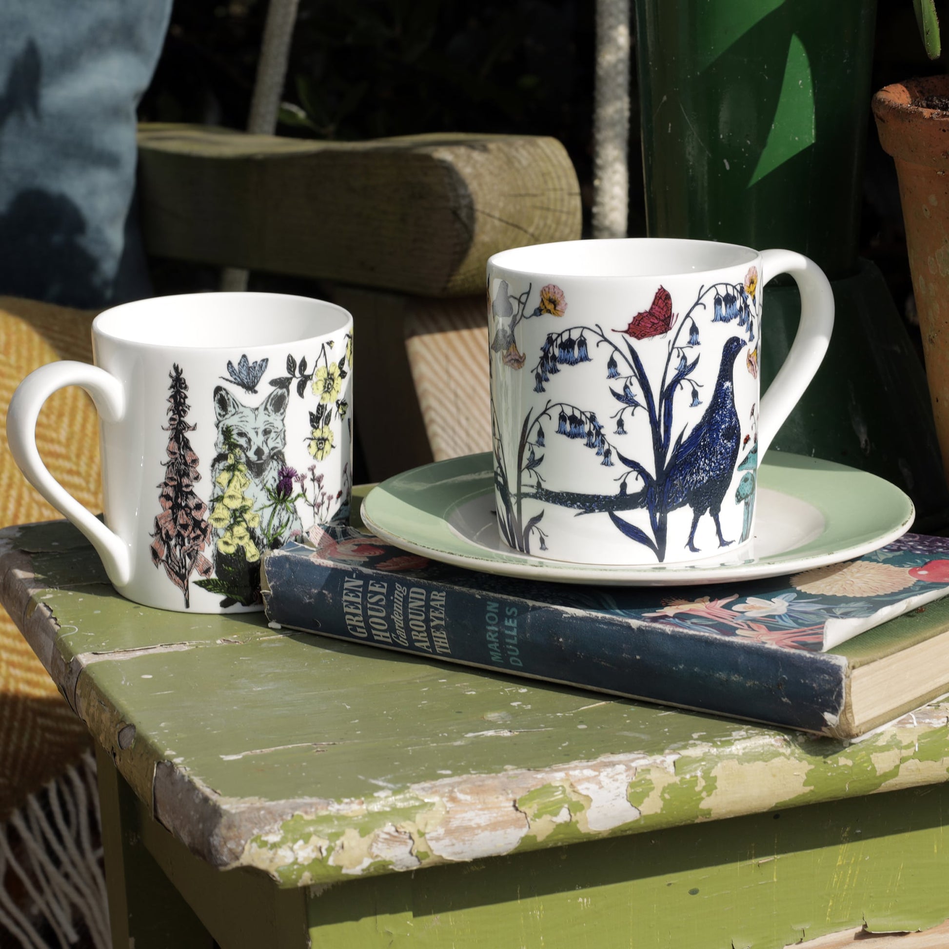Two illustrated bone china mugs, one with a fox and the other with a pheasant design, displayed on a weathered green table with vintage books, a plate, and garden decor.