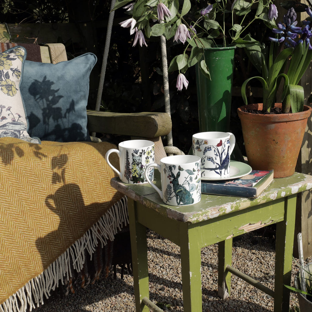 Three illustrated bone china mugs featuring a fox, hare, and pheasant displayed on a weathered green table in a garden setting, surrounded by potted plants, books, and a cosy bench with cushions and a mustard throw.