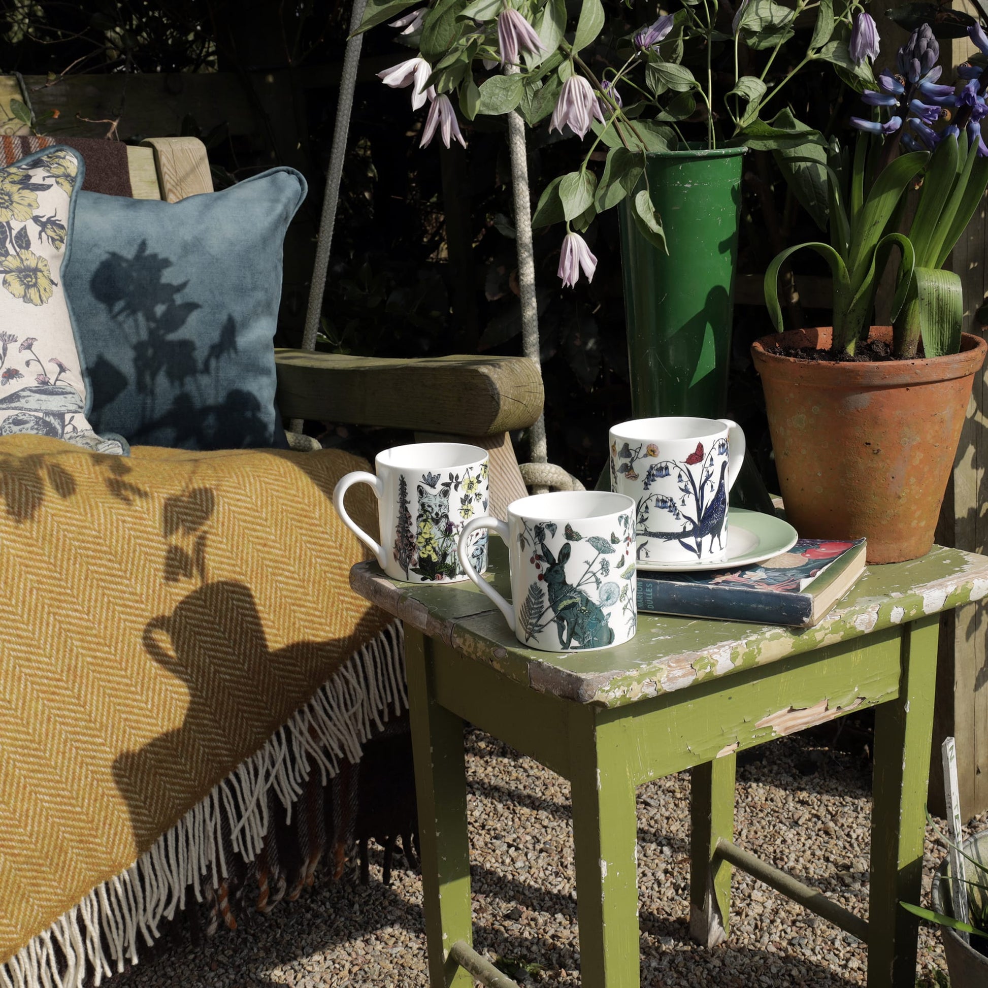Three illustrated bone china mugs featuring a fox, hare, and pheasant displayed on a weathered green table in a garden setting, surrounded by potted plants, books, and a cosy bench with cushions and a mustard throw.