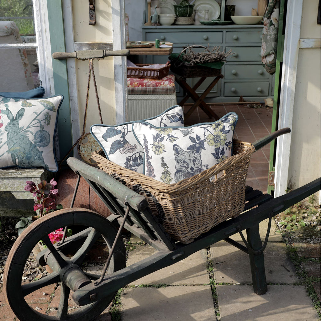 Rustic outdoor scene featuring a vintage wooden wheelbarrow with a woven basket filled with decorative cushions. The cushions display intricate illustrations of a fox and botanical elements. A charming countryside cottage setting with a pastel green kitchen dresser is visible in the background.