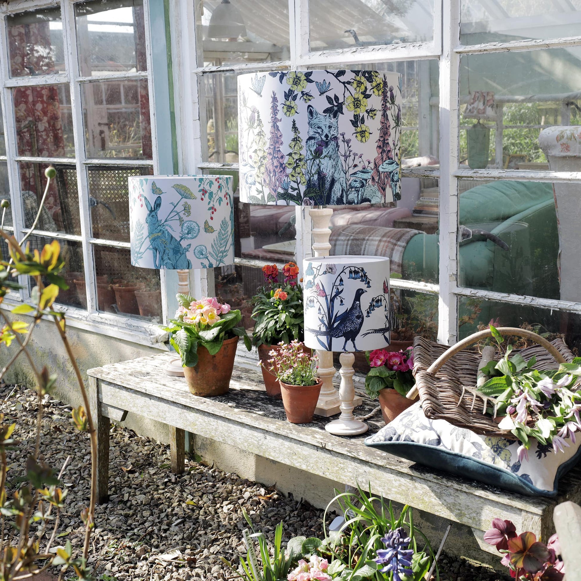 Rustic garden setting with a weathered wooden bench displaying three nature-inspired lampshades featuring a hare, fox, and pheasant among botanical details. Surrounding the lamps are potted flowers in terracotta pots, a wicker basket with freshly picked blooms, and a cushion with a matching design. A vintage greenhouse with aged window panes reflects the cozy interior beyond.