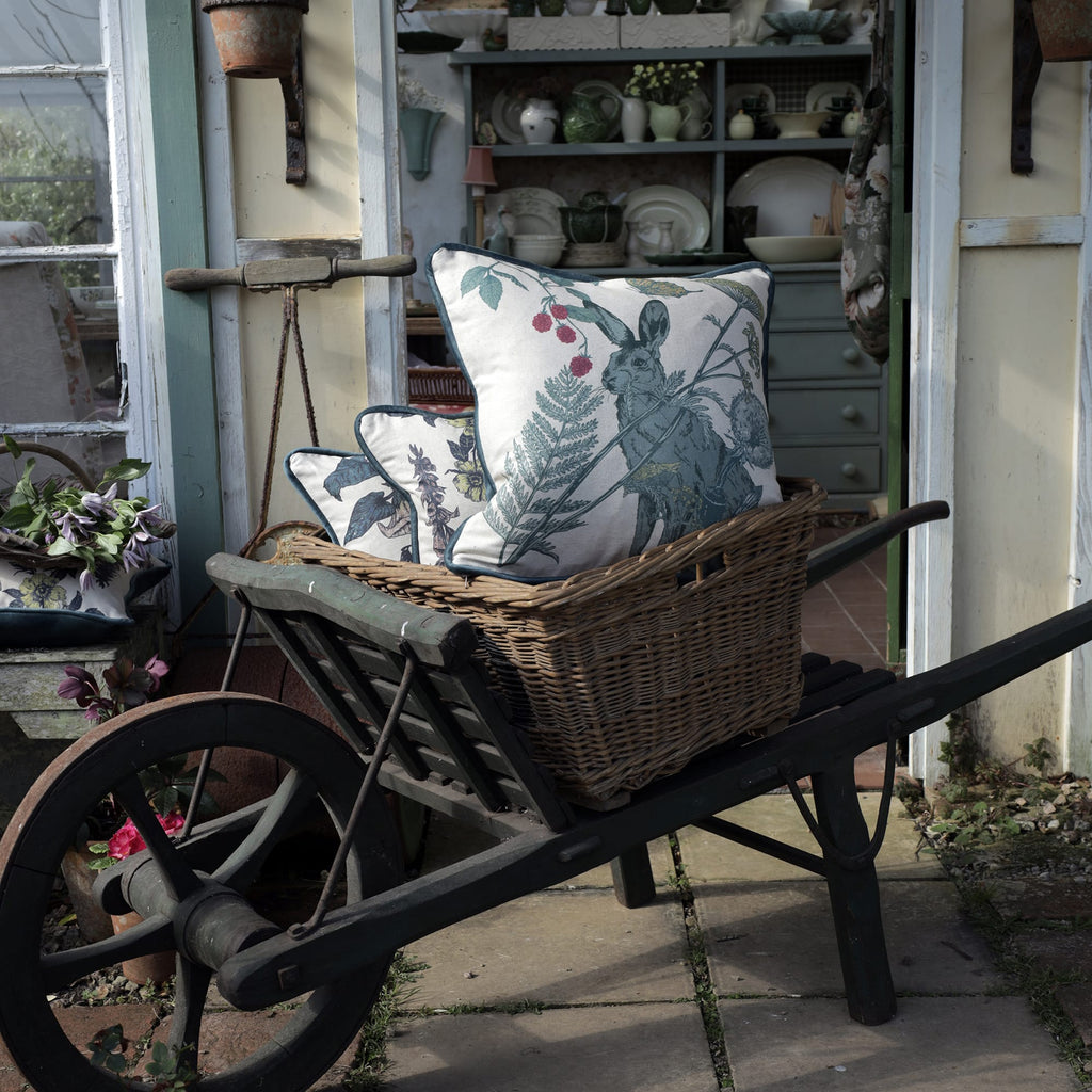 Vintage wooden wheelbarrow with a wicker basket, holding nature-inspired cushions featuring a hare and botanical designs. The setting is a rustic garden shed with an open door revealing a cozy interior filled with pastel green dishware, wooden furniture, and floral textiles, creating a charming countryside atmosphere.