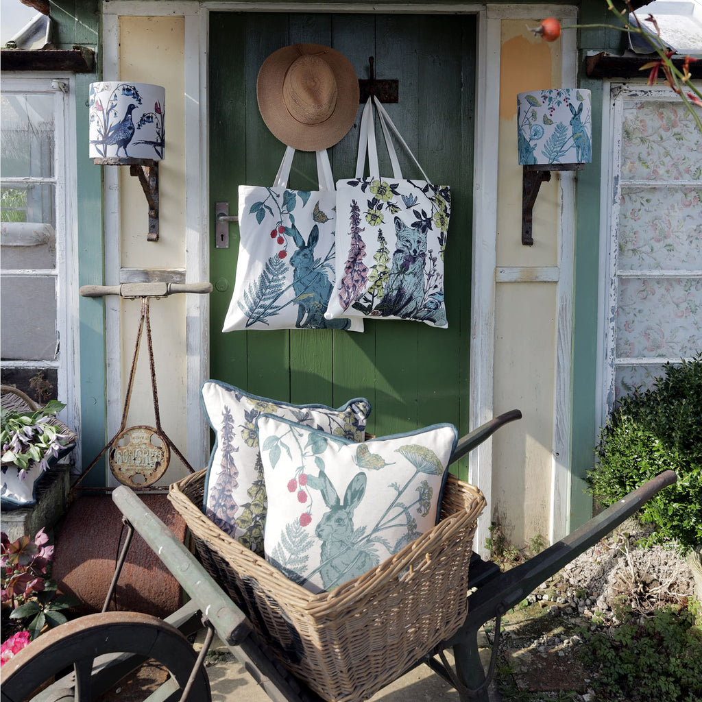 Charming cottage garden scene with a green wooden door adorned with two nature-themed tote bags and a straw hat. A vintage wheelbarrow with a wicker basket holds botanical-print cushions featuring a hare and wildflowers. Decorative lampshades with bird and foliage designs hang on either side, adding to the rustic countryside aesthetic.
