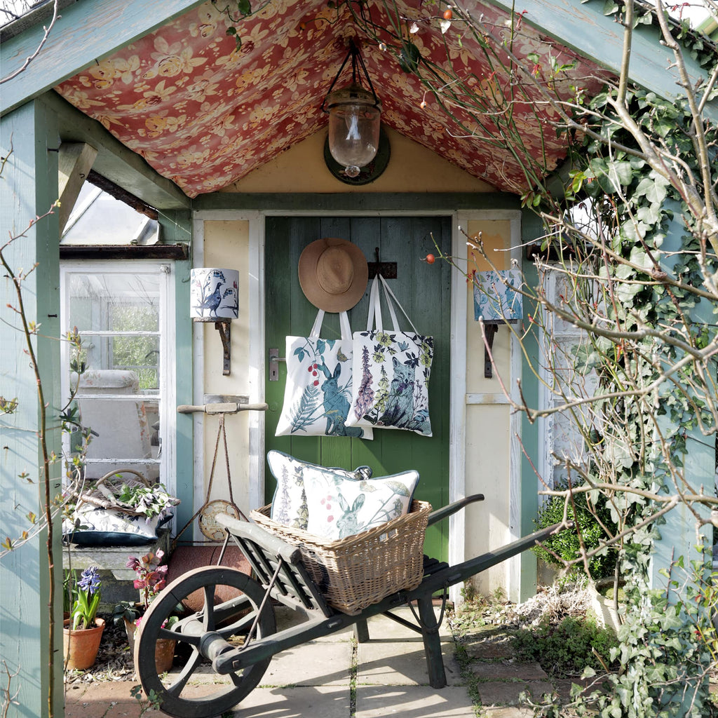 Rustic garden shed with a green door, decorated with two nature-themed tote bags, a straw hat, and a floral fabric ceiling. A vintage wheelbarrow with a woven basket holds a matching hare-print cushion. Potted plants, ivy, and a bird-print lamp add to the cozy countryside charm.