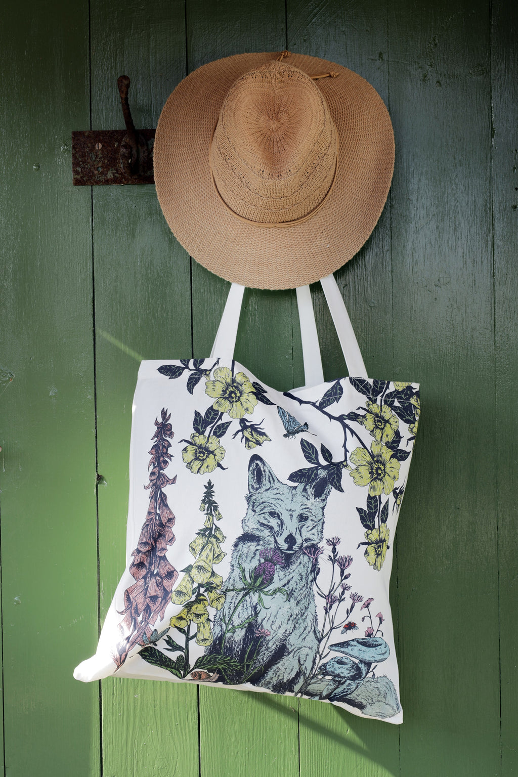 Nature-inspired tote bag featuring a detailed illustration of a fox surrounded by wildflowers and birds, hanging on a rustic green wooden door. A woven straw hat hangs above it on an antique metal hook, adding to the countryside charm. Sunlight casts soft shadows, highlighting the intricate design.