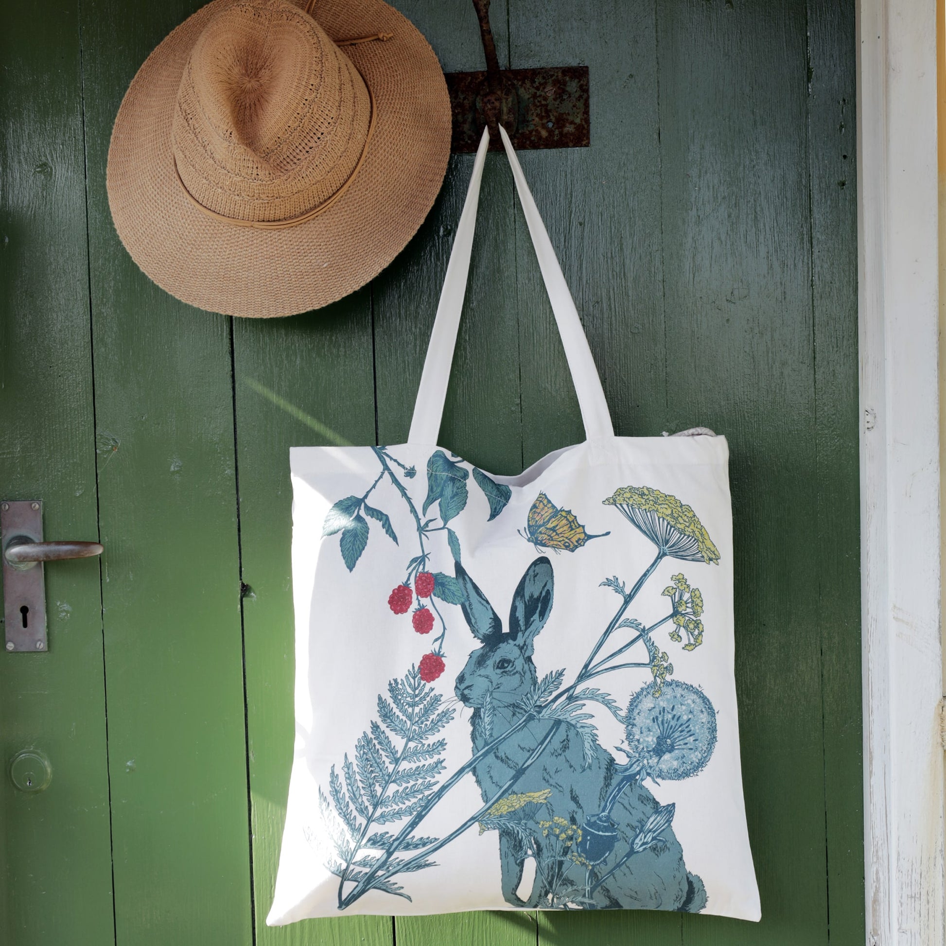 A white tote bag with an intricate nature-inspired design featuring a blue hare surrounded by wildflowers, ferns, raspberries, and a butterfly. The bag hangs on a rustic green wooden door alongside a woven straw hat, creating a countryside aesthetic with warm natural lighting.