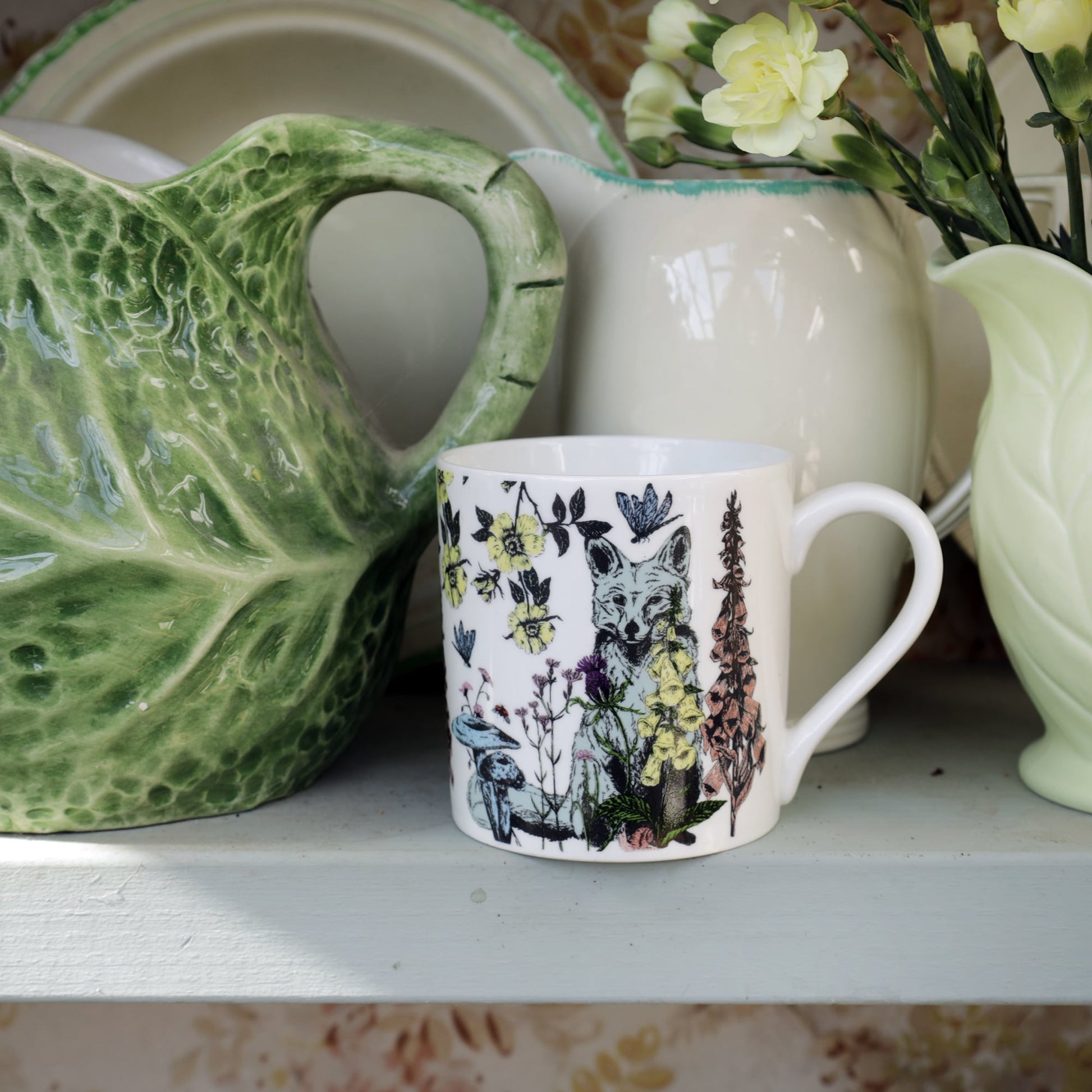 Bone china mug with a fox and wildflower illustration displayed on a shelf among vintage-style ceramic jugs, plates, and a vase of pale yellow flowers.