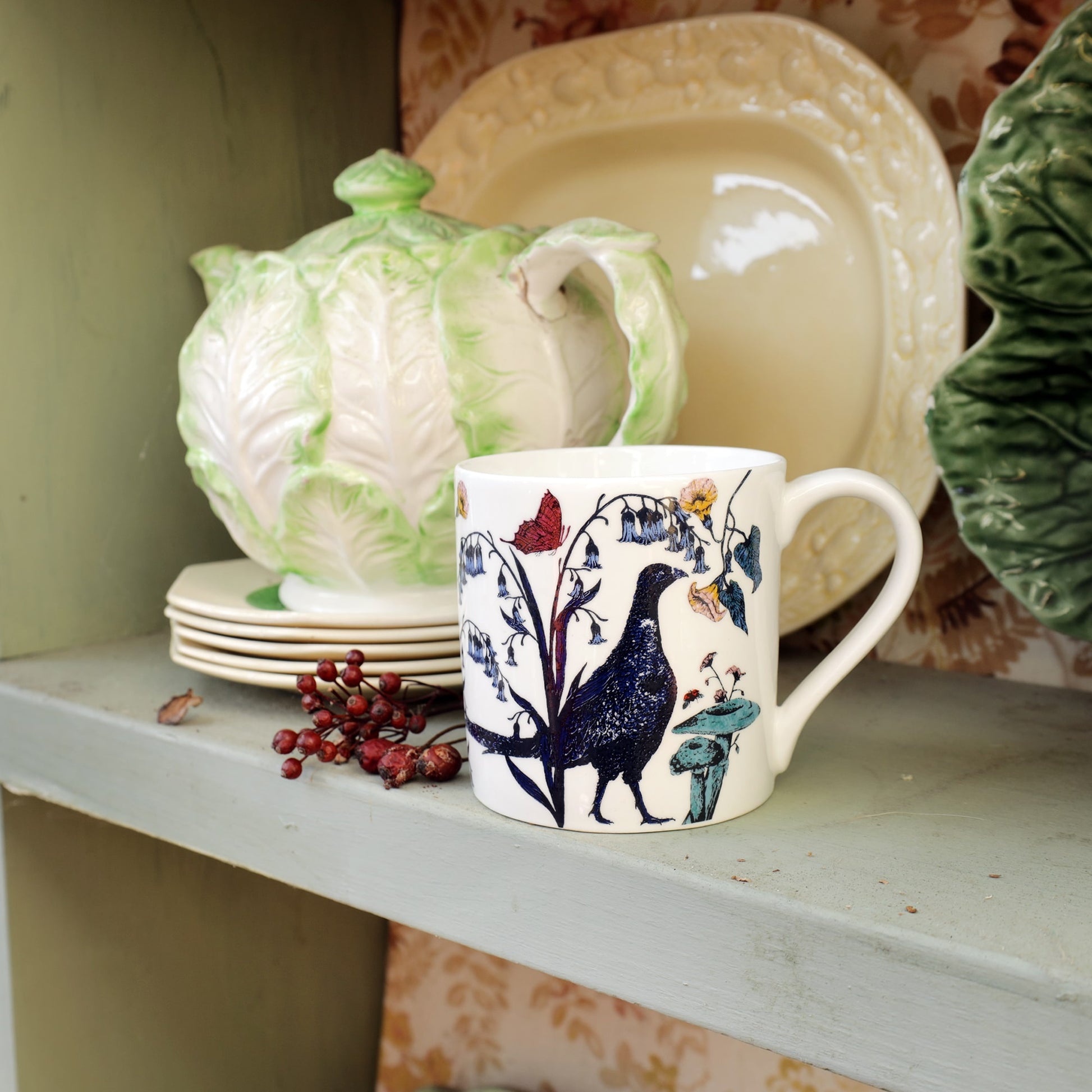 Bone china mug with a pheasant and wildflower illustration displayed on a vintage-style shelf alongside a cabbage-leaf teapot, stacked plates, and decorative ceramics.