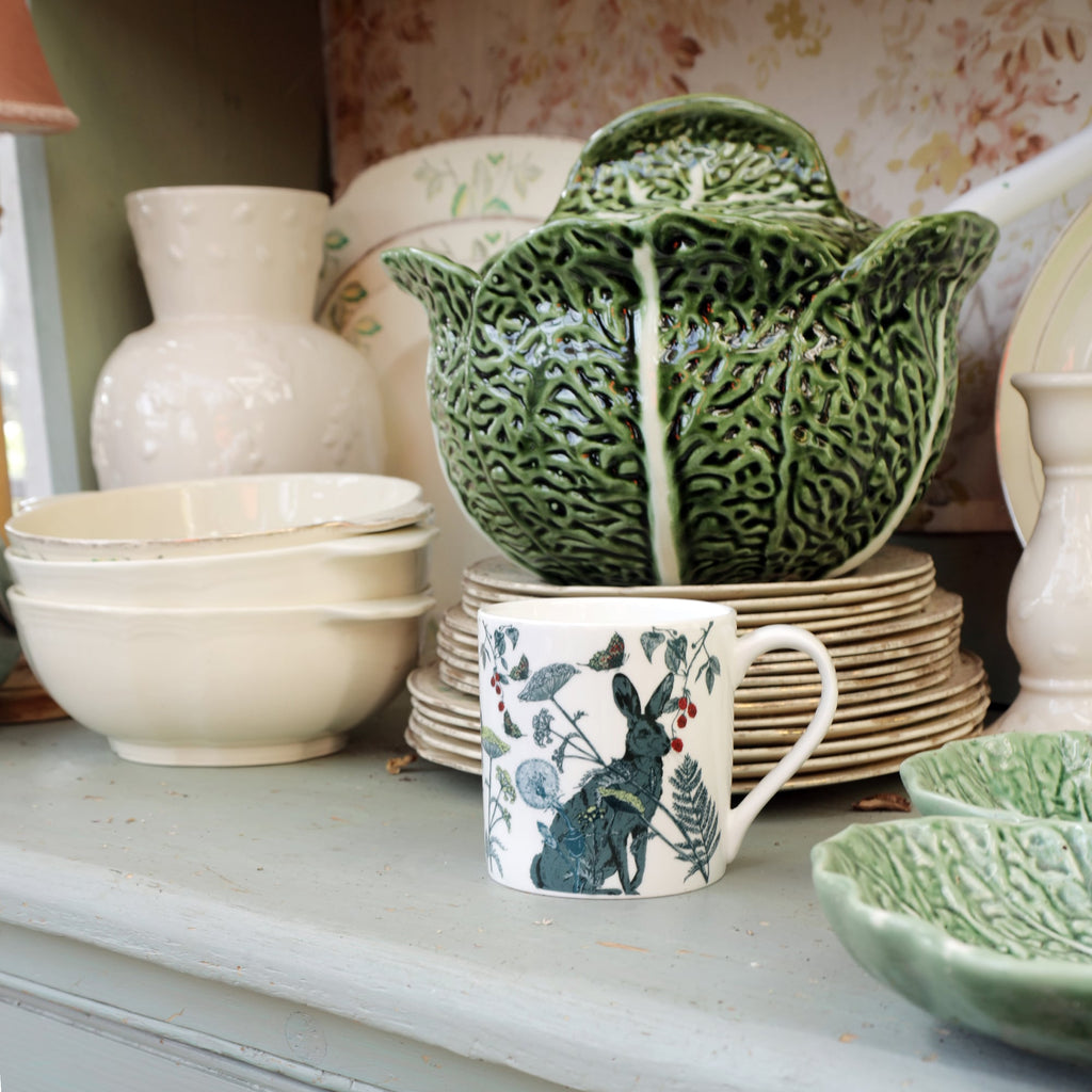 Bone china mug with a hare and botanical illustration displayed on a vintage shelf alongside stacked cream plates, bowls, and a green cabbage-leaf soup tureen.