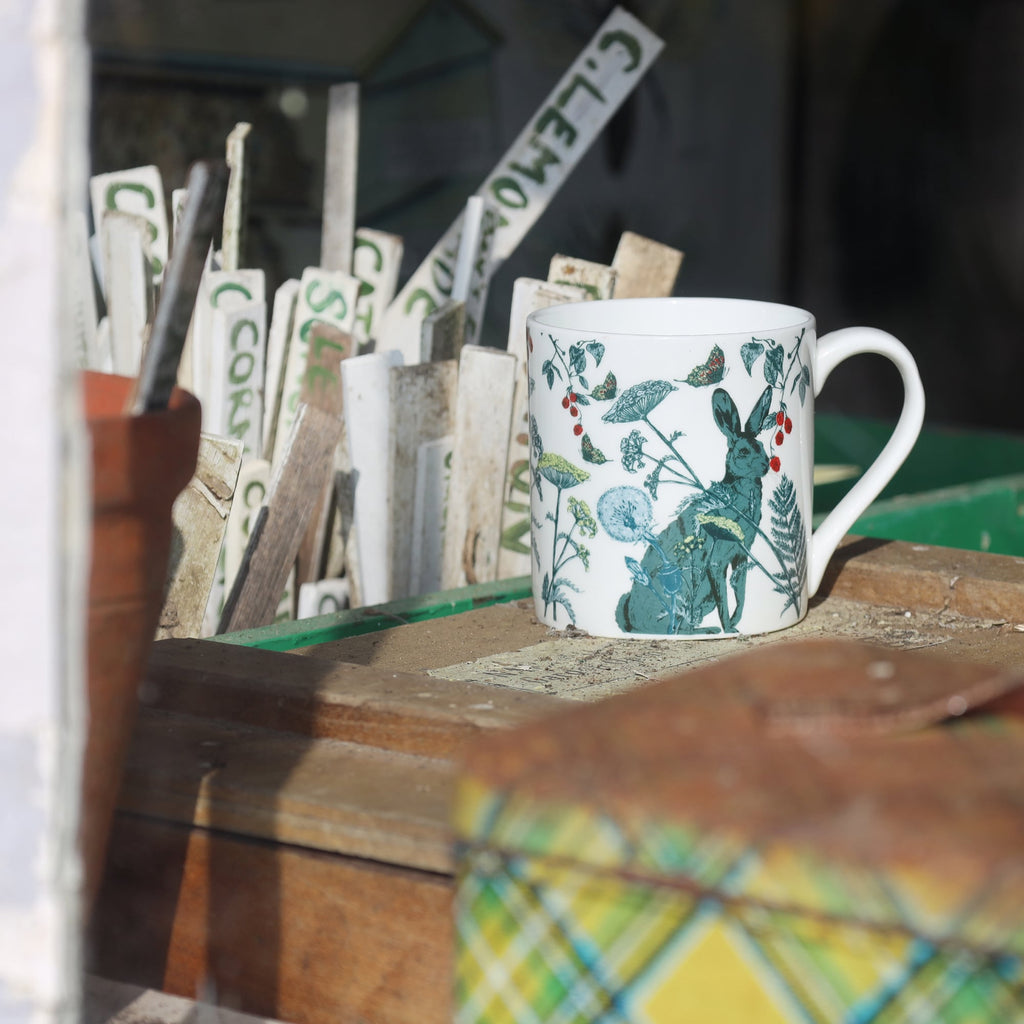 Bone china mug with a hare and wildflower illustration displayed on a rustic wooden surface in a potting shed, surrounded by vintage plant markers and garden tools.
