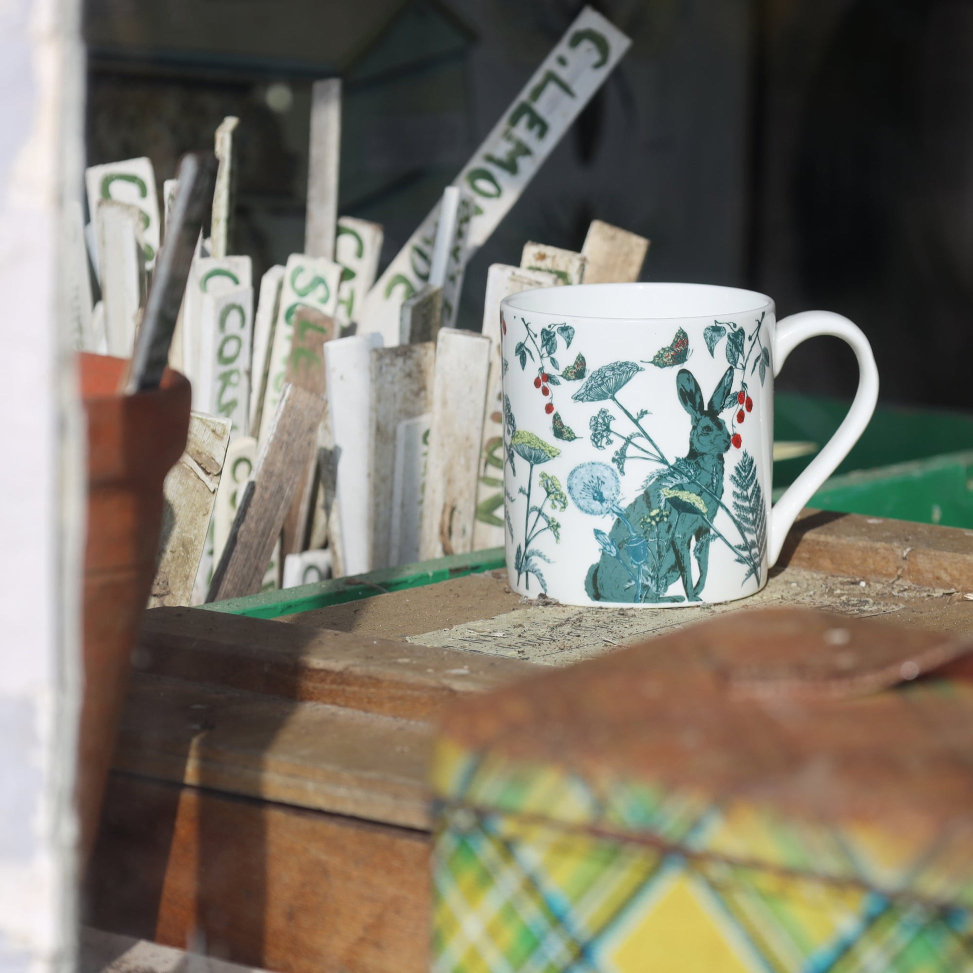 Bone china mug with a hare and wildflower illustration displayed on a rustic wooden surface in a potting shed, surrounded by vintage plant markers and garden tools.