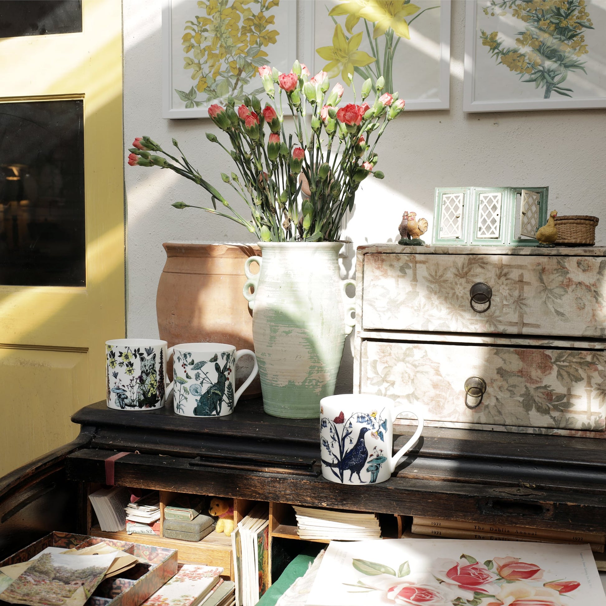 Three illustrated bone china mugs featuring a fox, hare, and pheasant arranged on a vintage writing desk beside floral prints, rustic pottery, and a vase of pink carnations in a sunlit room.