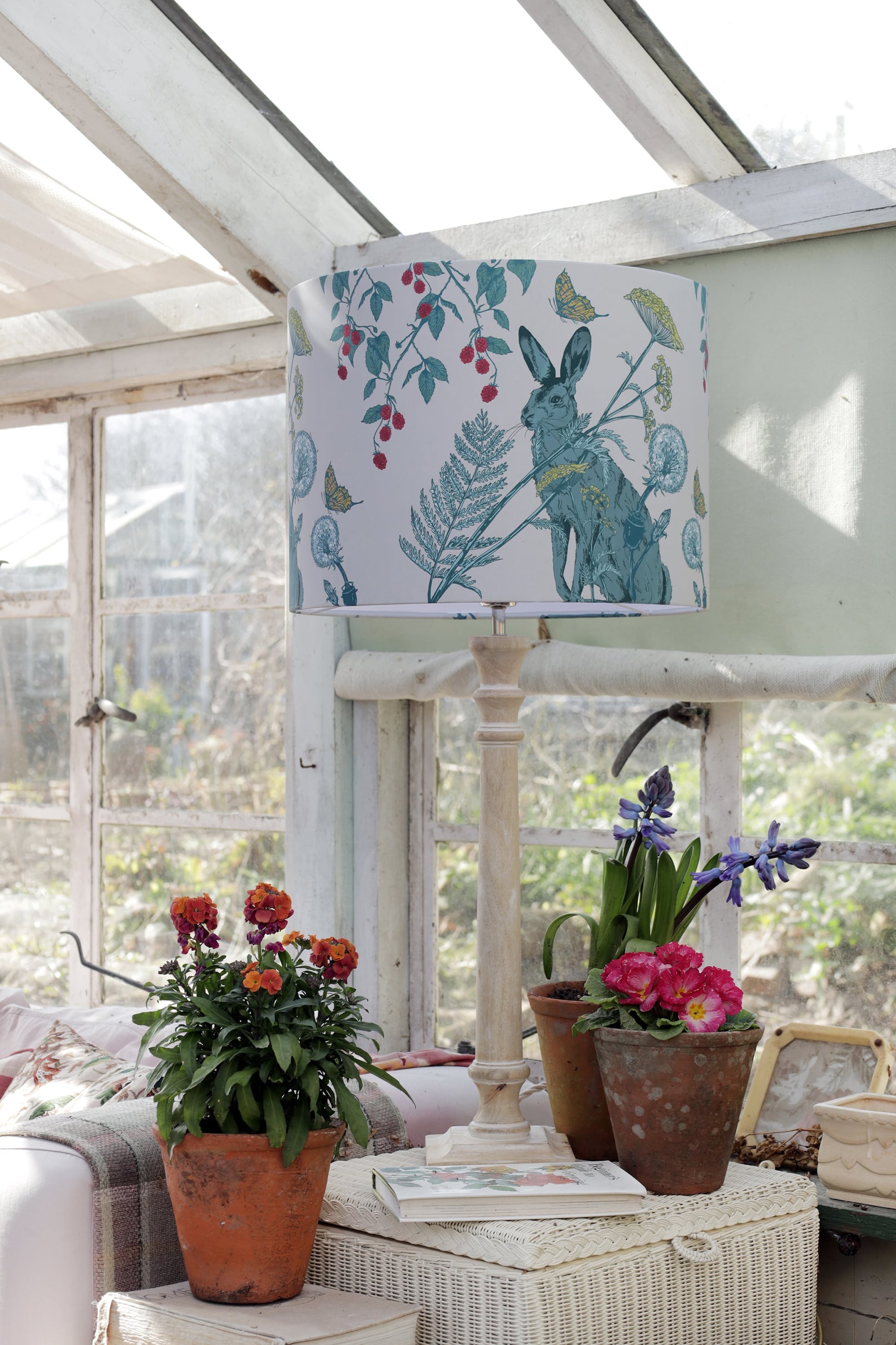 Charming greenhouse scene featuring a rabbit and berry print lampshade on a pale wooden lamp, surrounded by vibrant potted plants including primroses and hyacinths. The sun streams through the glass roof, casting light on the plants and a stack of books, creating a peaceful and inviting space.