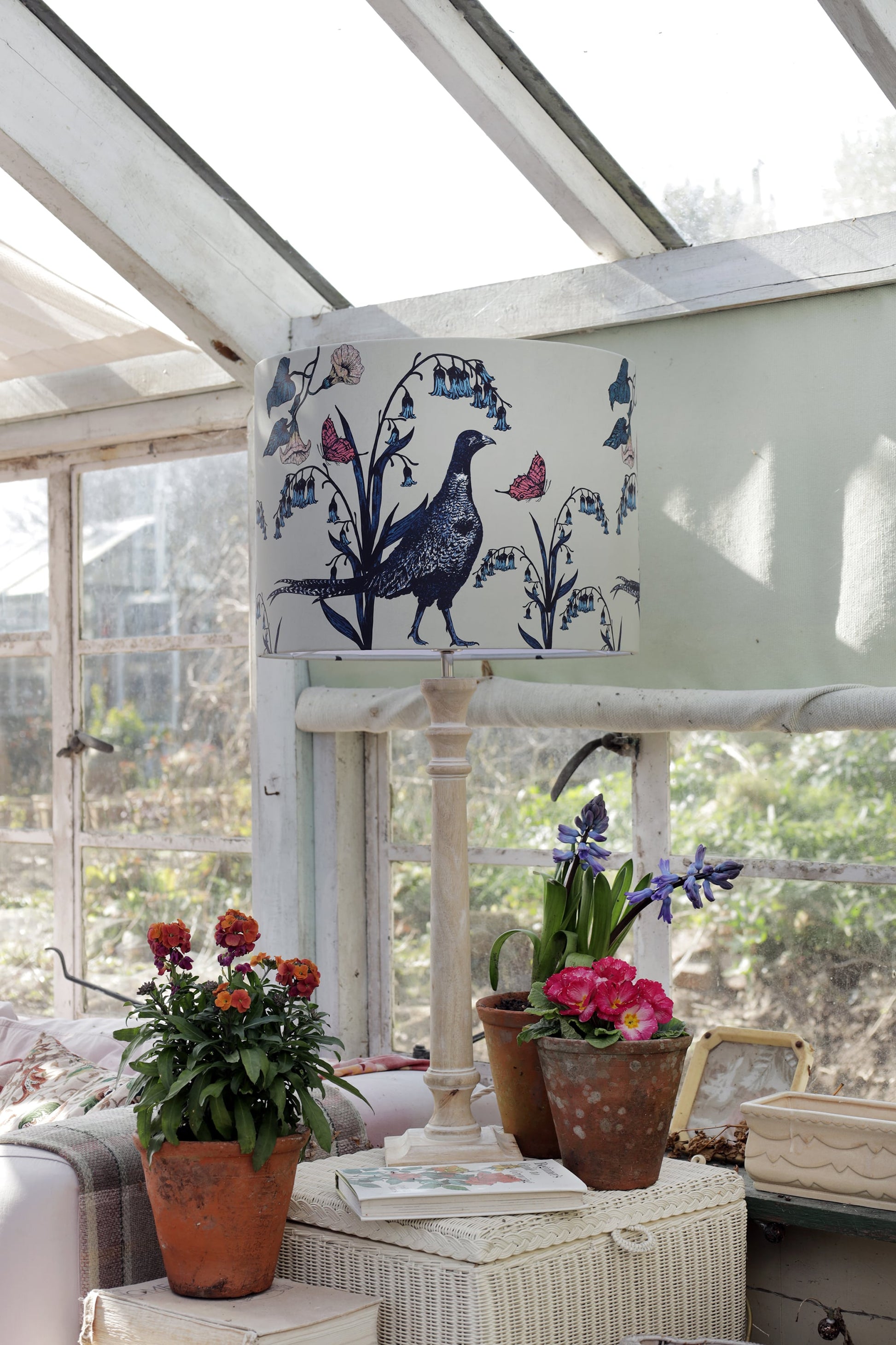 Sunlit greenhouse corner with a pheasant-themed lampshade featuring botanical details atop a wooden lamp base. Surrounding the lamp are terracotta pots filled with blooming flowers, a wicker storage box, and an open book, creating a cozy and rustic atmosphere with natural light streaming through vintage glass windows.