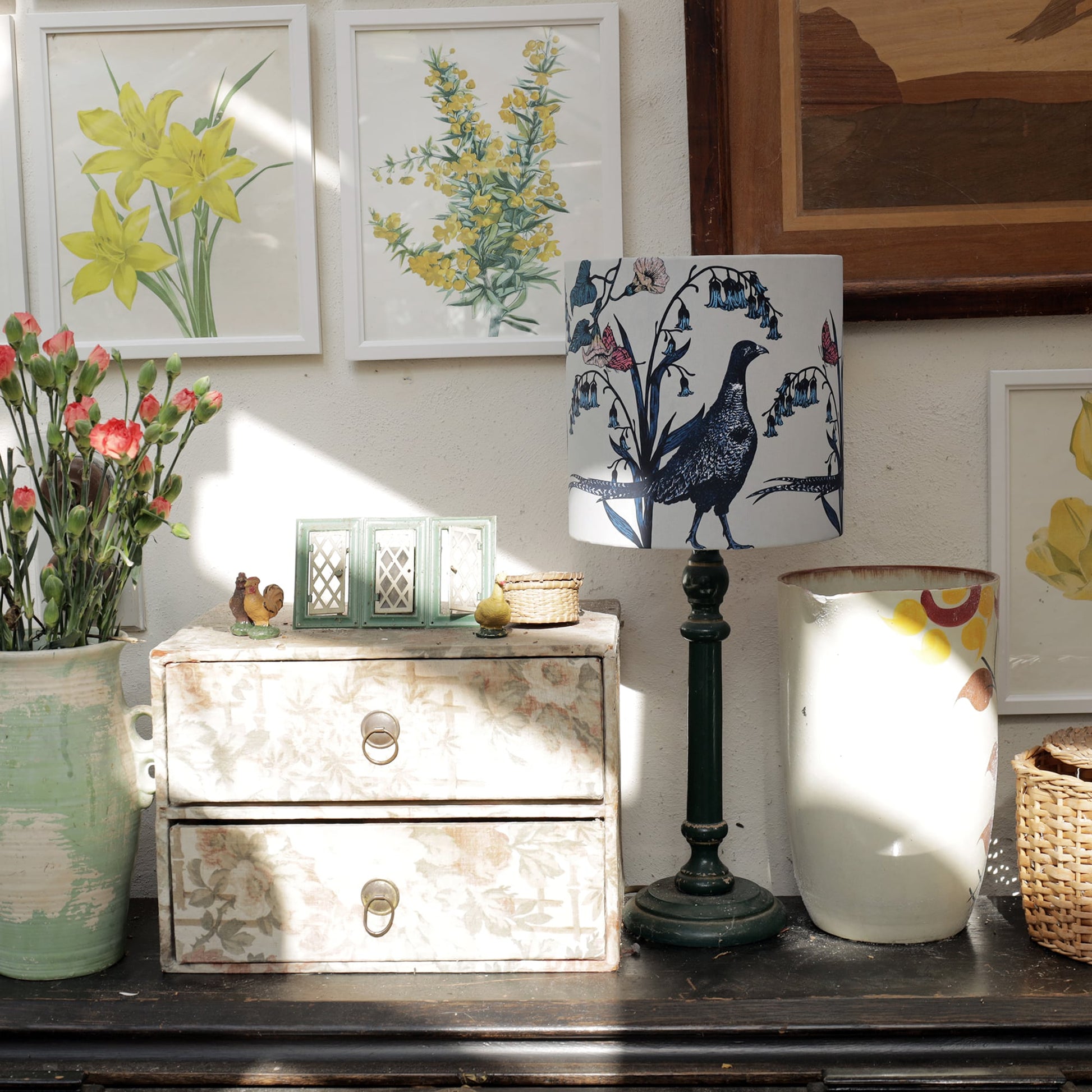 Sunlit interior with a dark wooden sideboard decorated with a pheasant-themed lampshade, botanical prints, and vintage floral storage drawers. A ceramic pitcher with pink flowers, a wicker basket, and small decorative accents add to the rustic charm, while framed yellow floral artwork brightens the space.