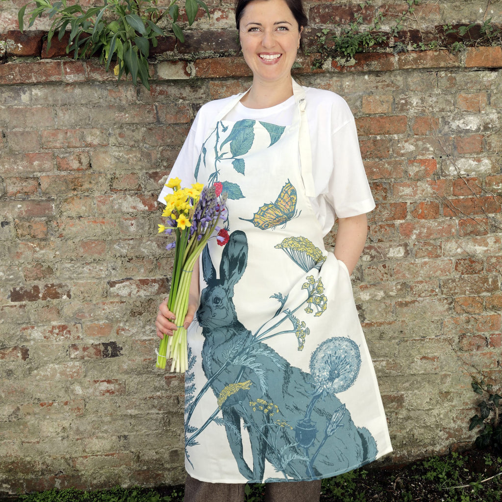 Smiling woman standing in front of a brick wall, wearing a white t-shirt and an apron featuring an illustrated hare surrounded by botanical elements like flowers, berries, and butterflies. She is holding a bouquet of spring flowers in one hand.