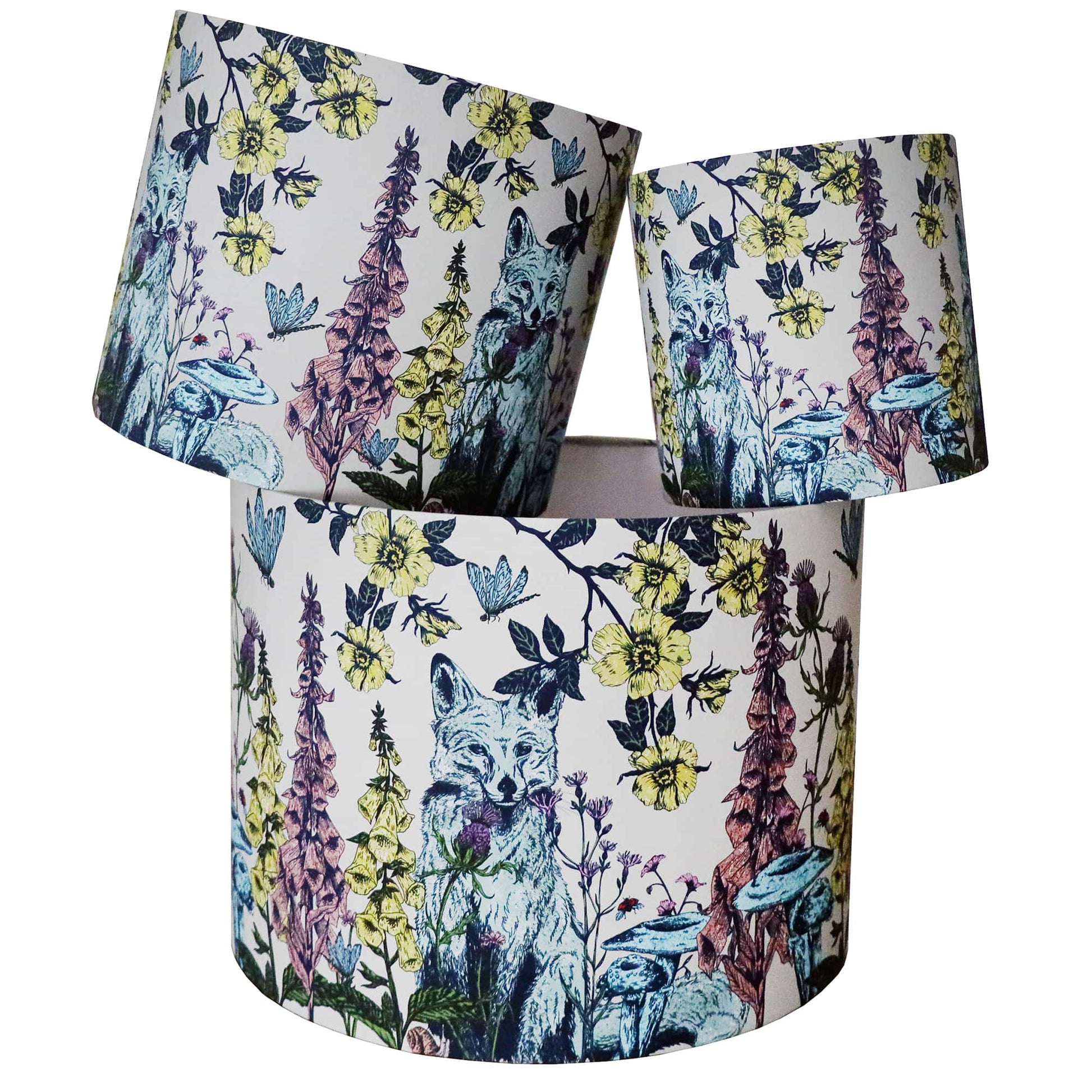 A set of three lampshades featuring an intricate woodland-inspired design with a fox surrounded by wildflowers, mushrooms, and buzzing insects. The artwork is detailed with soft blues, yellows, and earthy tones on a cream background, evoking a charming countryside aesthetic. The lampshades are stacked at different angles, highlighting the full pattern and the nature-inspired elements.