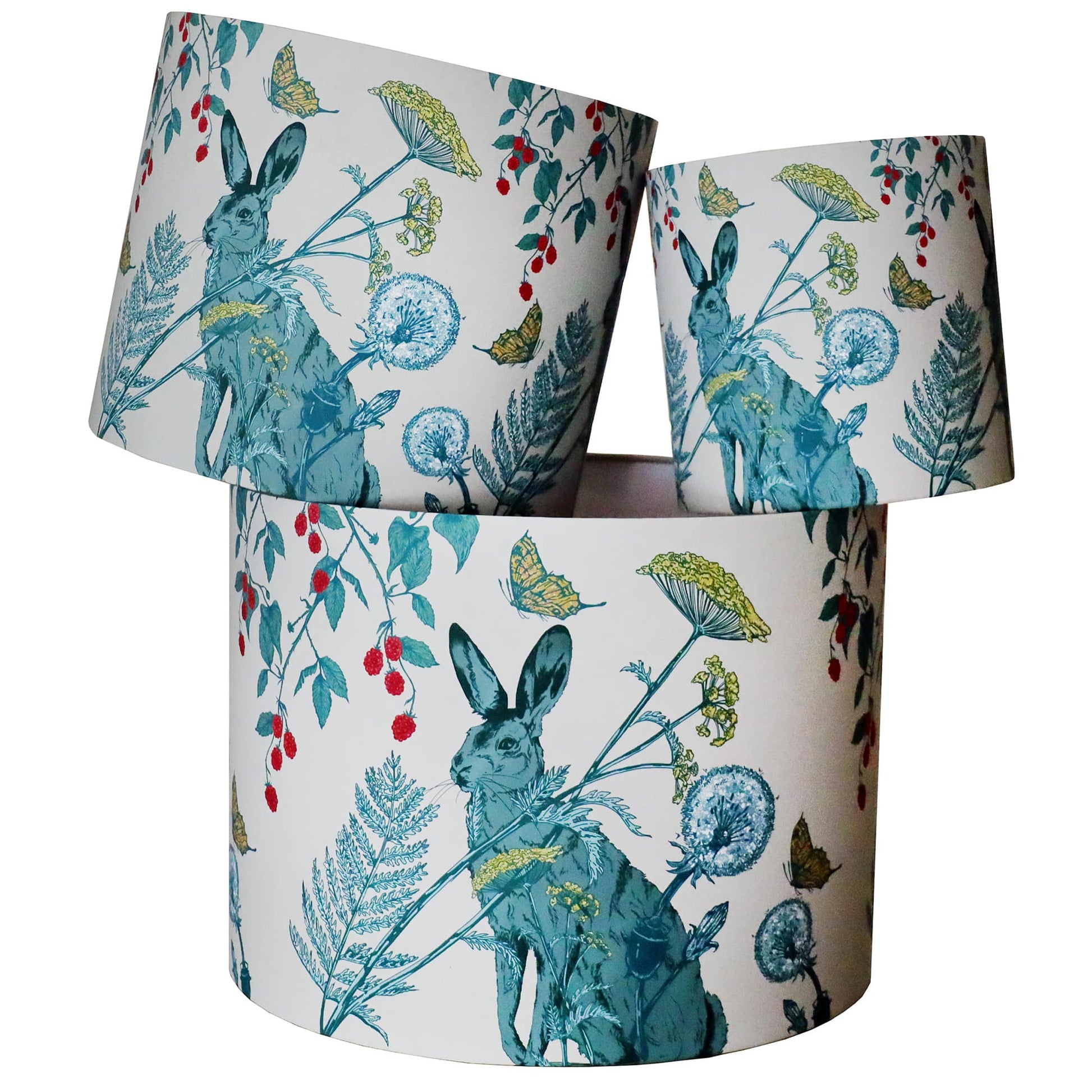 A set of three fabric lampshades featuring a beautifully illustrated hare surrounded by wildflowers, dandelions, berries, and butterflies. The detailed design is rendered in soft blues, greens, and golden yellows on a cream background, evoking a whimsical countryside feel. The lampshades are stacked to showcase different angles of the nature-inspired pattern, creating an eye-catching and elegant display.