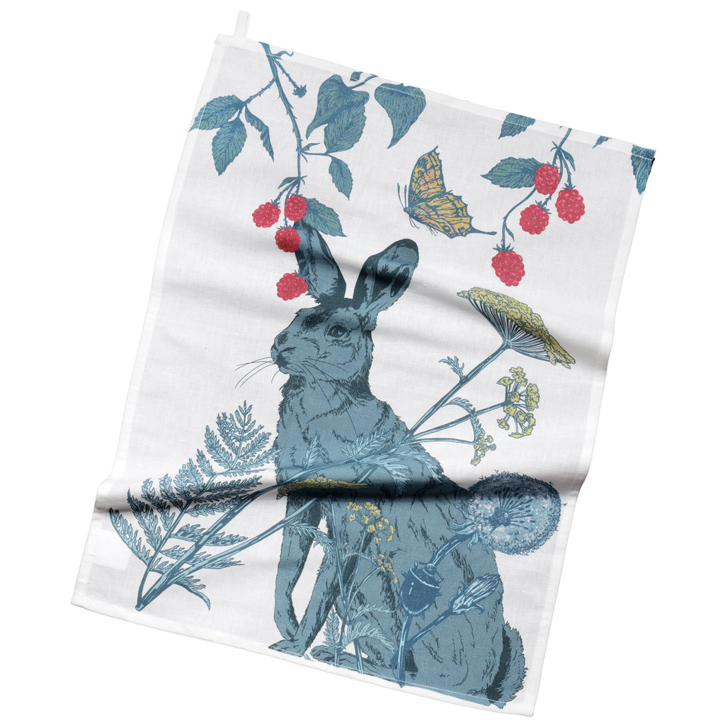 Tea towel featuring a detailed illustration of a hare surrounded by wildflowers, ferns, dandelions, and raspberry vines. A butterfly hovers above, adding a whimsical touch to the nature-inspired design in shades of green, red, and yellow on a white background.