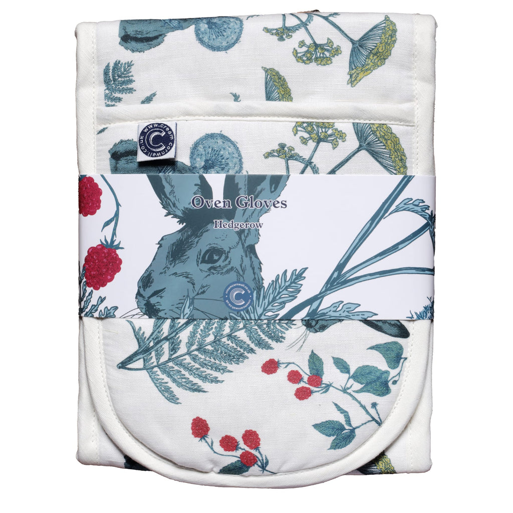 Packaged double oven gloves with a countryside-inspired print of a hare surrounded by wildflowers, ferns, dandelions, and raspberry vines. The gloves are wrapped in a branded label that reads 'Oven Gloves – Hedgerow,' showcasing the rustic nature-themed design in shades of green, red, and yellow on a white background