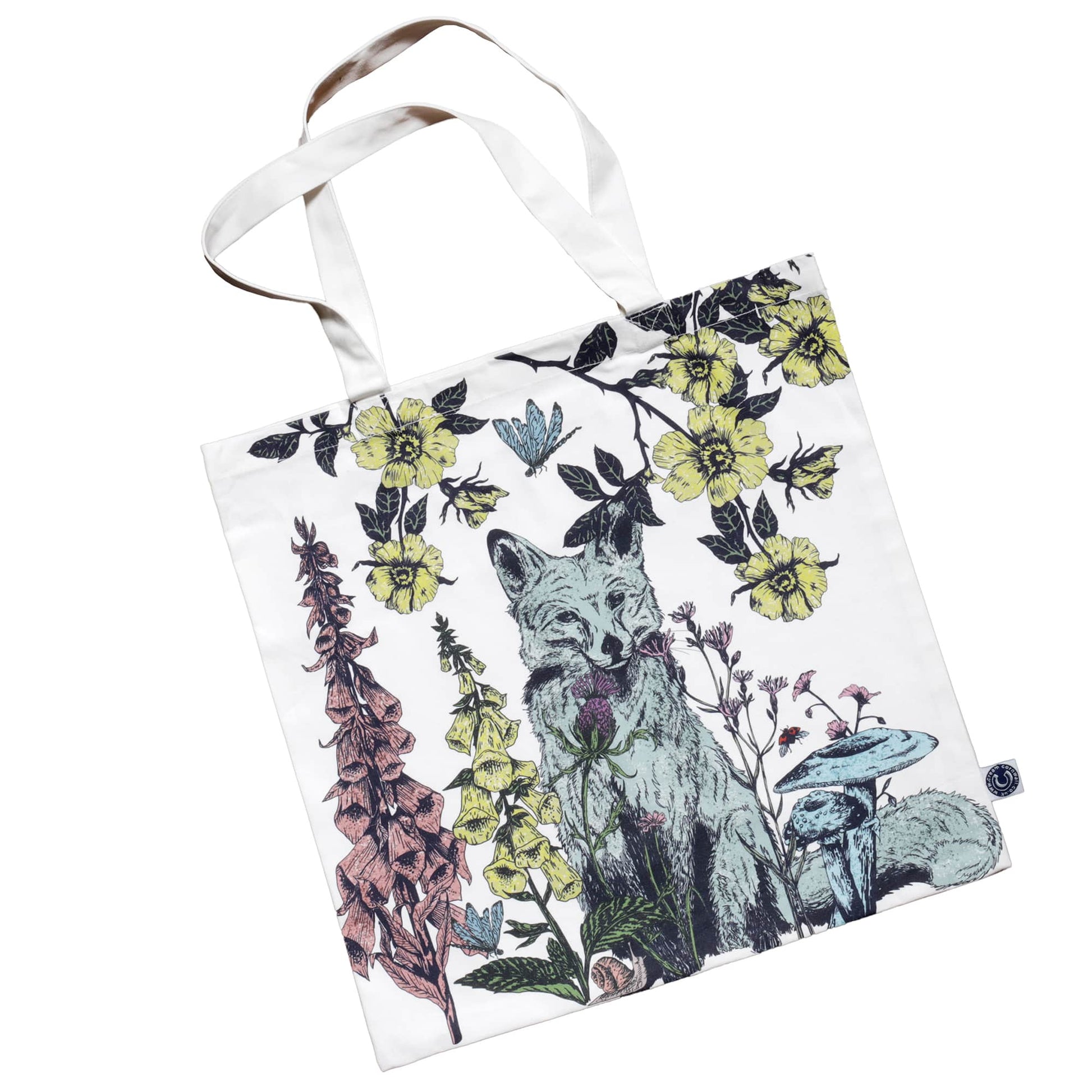 Fabric tote bag featuring a woodland-themed illustration of a fox surrounded by foxgloves, yellow wildflowers, mushrooms, and climbing vines. The design includes a butterfly and a ladybird, with a nature-inspired color palette of green, yellow, pink, and blue on a white background. The bag has sturdy white handles.