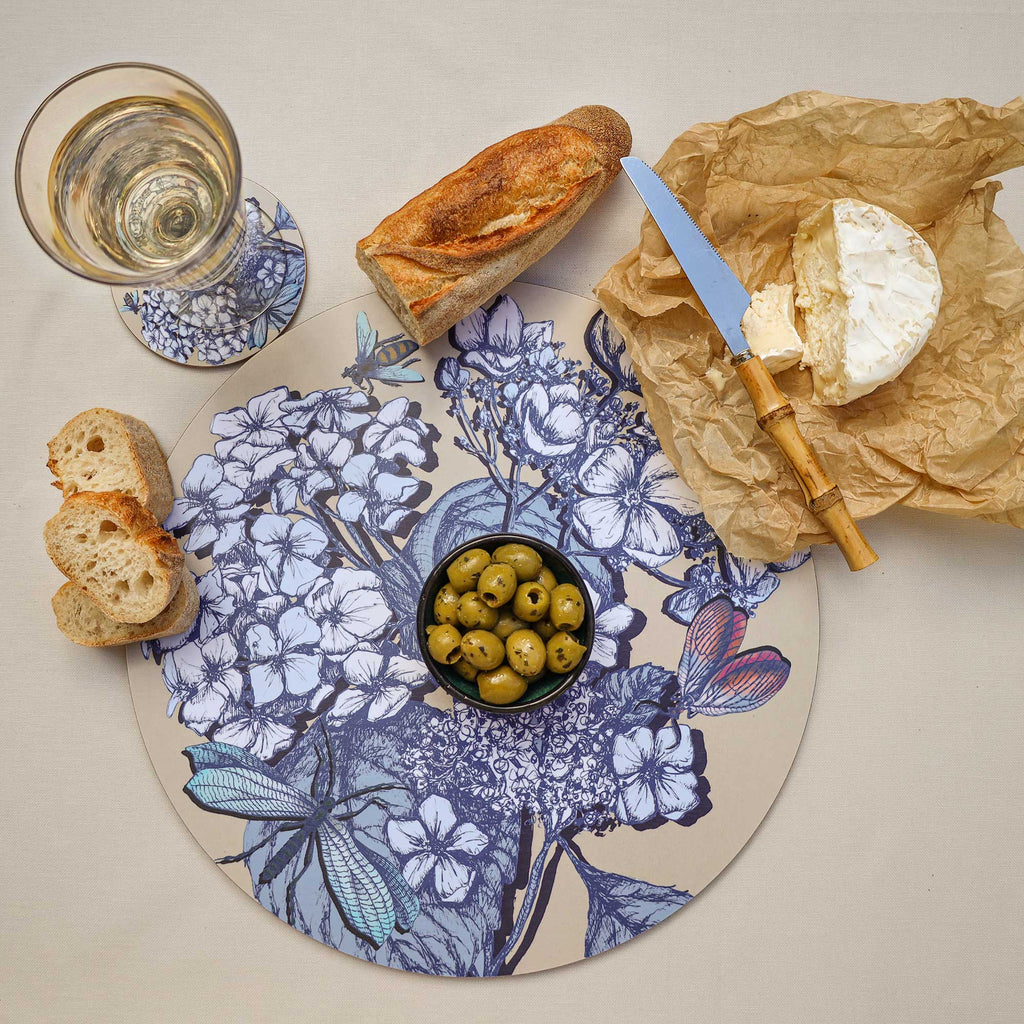 
Overhead view of a round cream placemat featuring intricate blue and white botanical illustrations of flowers, leaves, a turquoise dragonfly, a bee and a colourful butterfly. Styled on a neutral linen tablecloth with a bowl of green olives, crusty baguette, sliced bread, soft cheese on brown parchment paper, a bamboo-handled cheese knife and a glass of white wine resting on a matching floral coaster.