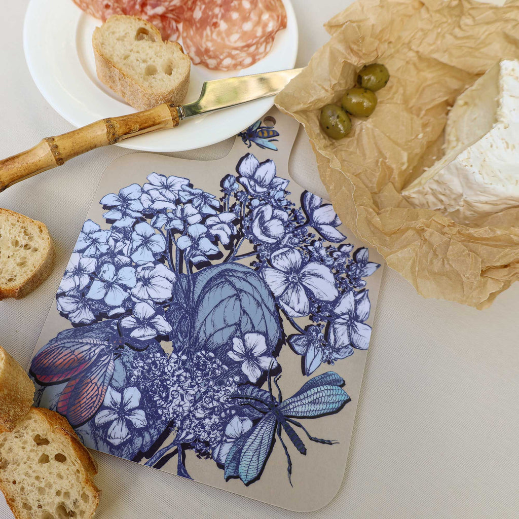 Cream Cornwall hydrangea chopping board featuring intricate blue botanical illustrations, butterflies and dragonflies, styled with creamy Brie cheese, green olives, sliced salami and crusty bread on a neutral linen tablecloth.