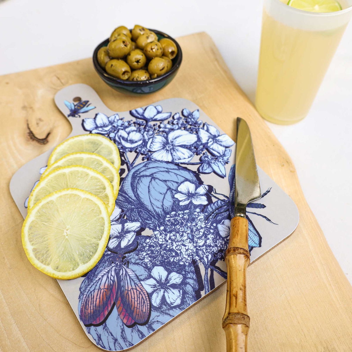 
Cream Cornwall hydrangea chopping board featuring intricate blue botanical illustrations and colourful dragonflies, styled with fresh lemon slices, a bamboo-handled knife, a bowl of green olives and a refreshing citrus drink on a natural wooden surface.