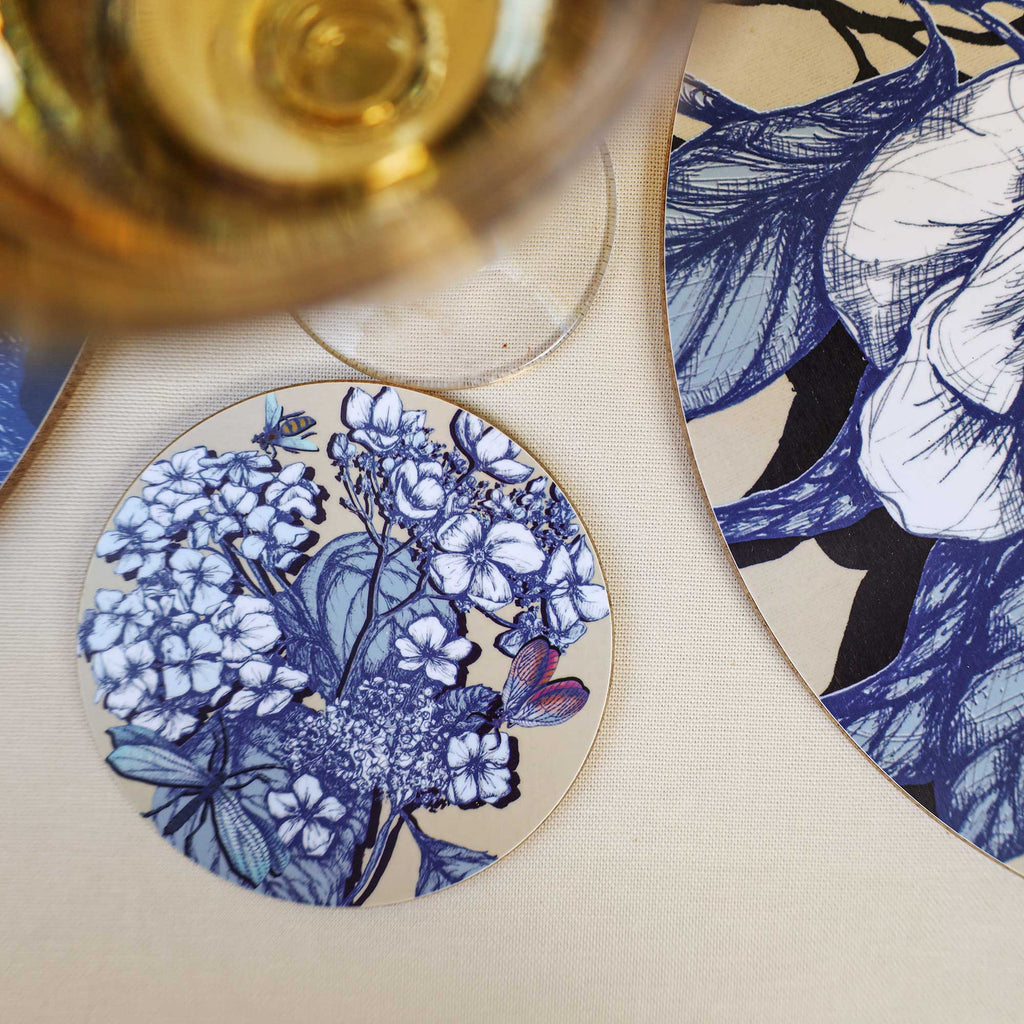 Cream Cornwall round hydrangea coaster featuring intricate blue and white botanical illustrations, delicate butterflies, a dragonfly and a bee on a soft cream background, styled beside a matching floral placemat and a glass of white wine on a neutral linen tablecloth.