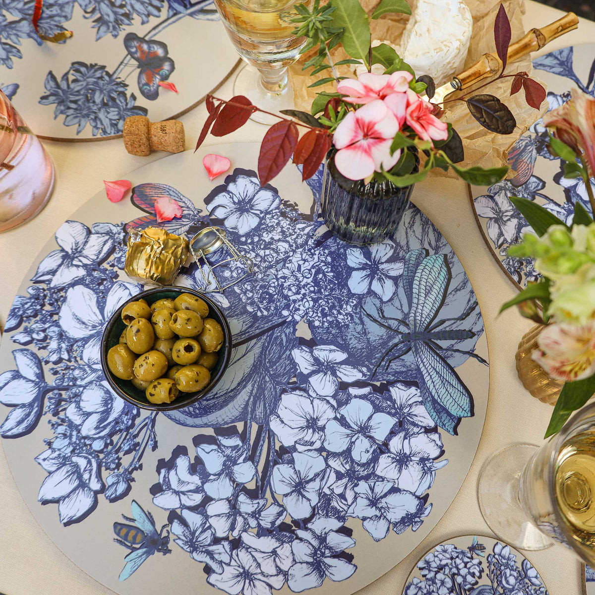 
Overhead view of a Cream Cornwall round placemat featuring intricate blue and white botanical illustrations, delicate flowers, butterflies, bees and a large dragonfly on a soft cream background. Styled with a bowl of green olives, pink flowers in a dark glass vase, a glass of white wine, soft cheese on parchment paper, bamboo-handled cutlery and matching botanical tableware on a neutral linen tablecloth.