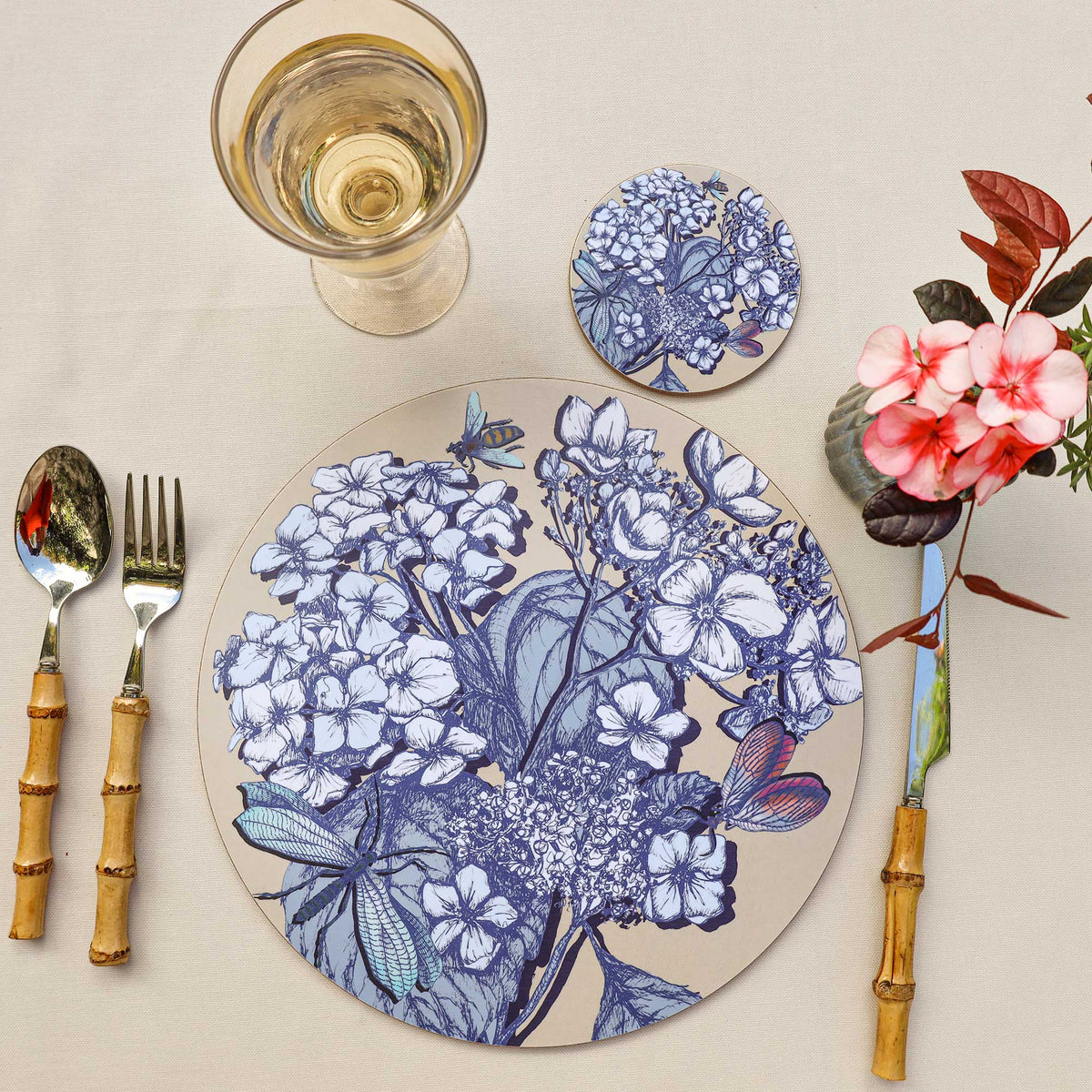 
Overhead view of an elegant table setting featuring a round placemat with intricate blue botanical illustrations of hydrangeas, leaves, dragonflies, a bee and a colourful butterfly on a beige background. A matching floral coaster sits above the placemat, accompanied by a glass of white wine, bamboo-handled cutlery and a small arrangement of pink flowers with burgundy leaves, all styled on a cream linen tablecloth.