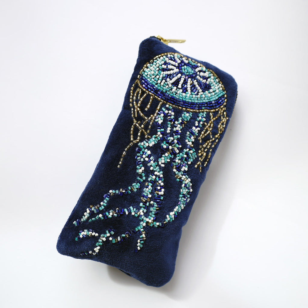 A navy blue velvet glasses case featuring an intricate beaded jellyfish design. The jellyfish is crafted with shimmering beads in shades of turquoise, white, gold, and deep blue, with flowing tentacles trailing down the length of the case. The case has a soft, padded texture and a gold zipper at the top, set against a clean white background.