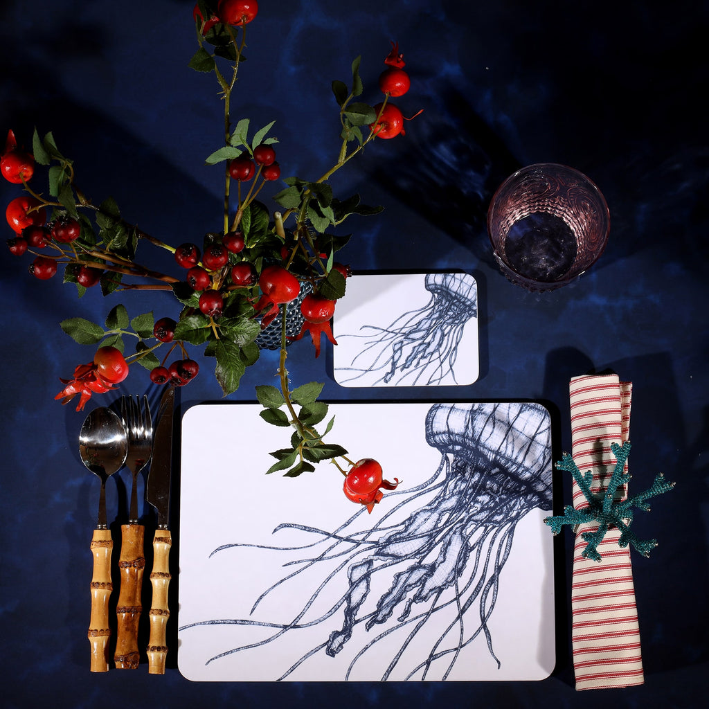 Jellyfish Design in Navy on a white Coaster with a matching Placemat in a table setting on a dark table cloth.On the table is bamboo cutlery, stripe napkin and a glass with rosehips.