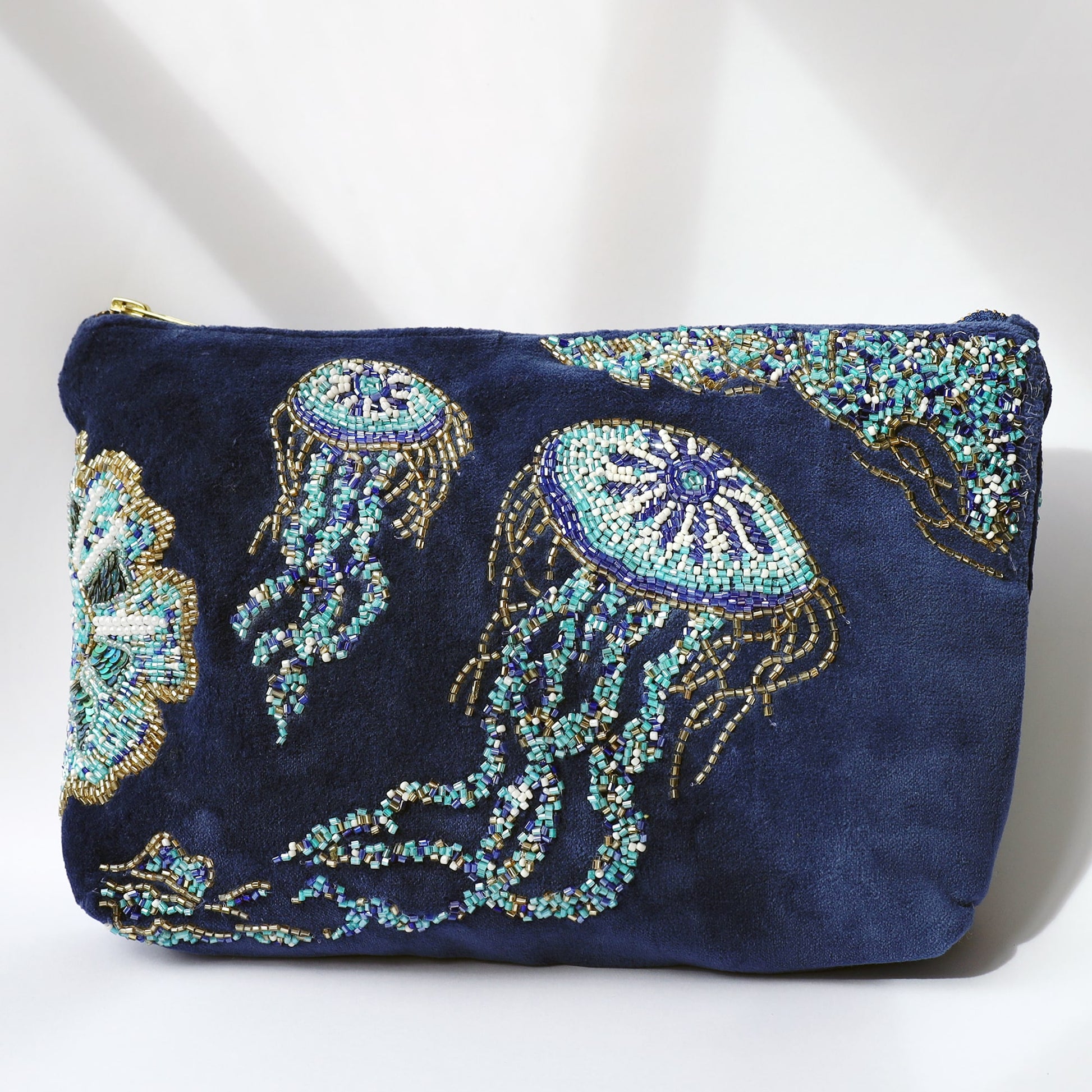 A deep navy velvet pouch adorned with intricate bead embroidery of jellyfish in shimmering shades of turquoise, gold, and white. The jellyfish designs feature flowing tentacles and circular tops, creating a dynamic underwater scene. The pouch has a gold zip closure and is photographed under soft natural lighting that highlights the texture and sparkle of the beads.