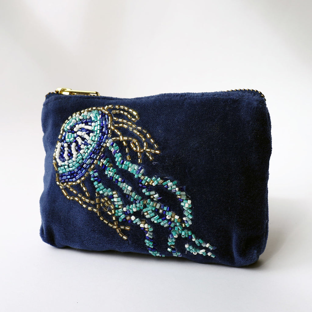 A small navy velvet purse featuring a detailed beaded jellyfish design in shades of turquoise, white, blue, and gold. The jellyfish is intricately embroidered, with long trailing tentacles and a rounded bell. The purse has a gold zip closure at the top and is photographed against a light background, highlighting the rich texture and shimmering beadwork.