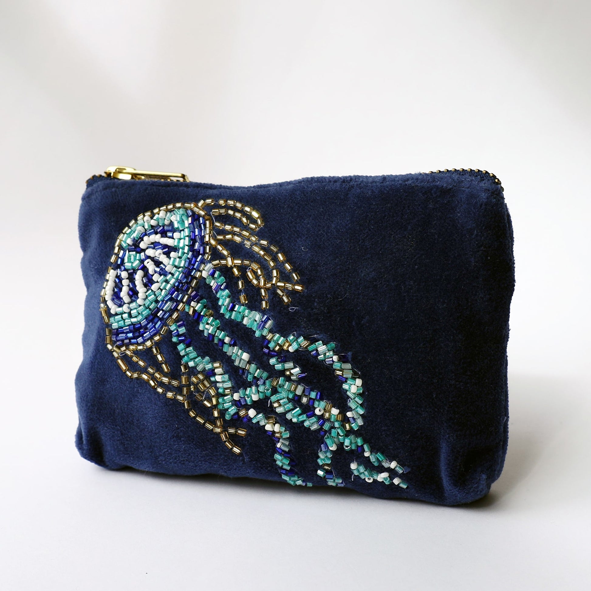 A small navy velvet purse featuring a detailed beaded jellyfish design in shades of turquoise, white, blue, and gold. The jellyfish is intricately embroidered, with long trailing tentacles and a rounded bell. The purse has a gold zip closure at the top and is photographed against a light background, highlighting the rich texture and shimmering beadwork.