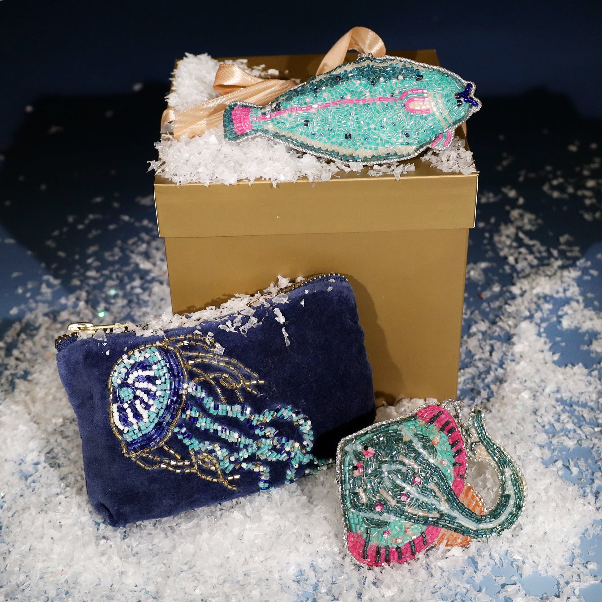 Navy velvet coin purse with an intricate beaded jellyfish design, displayed beside a gold gift box topped with a turquoise beaded fish ornament and surrounded by faux snow for a festive coastal look.