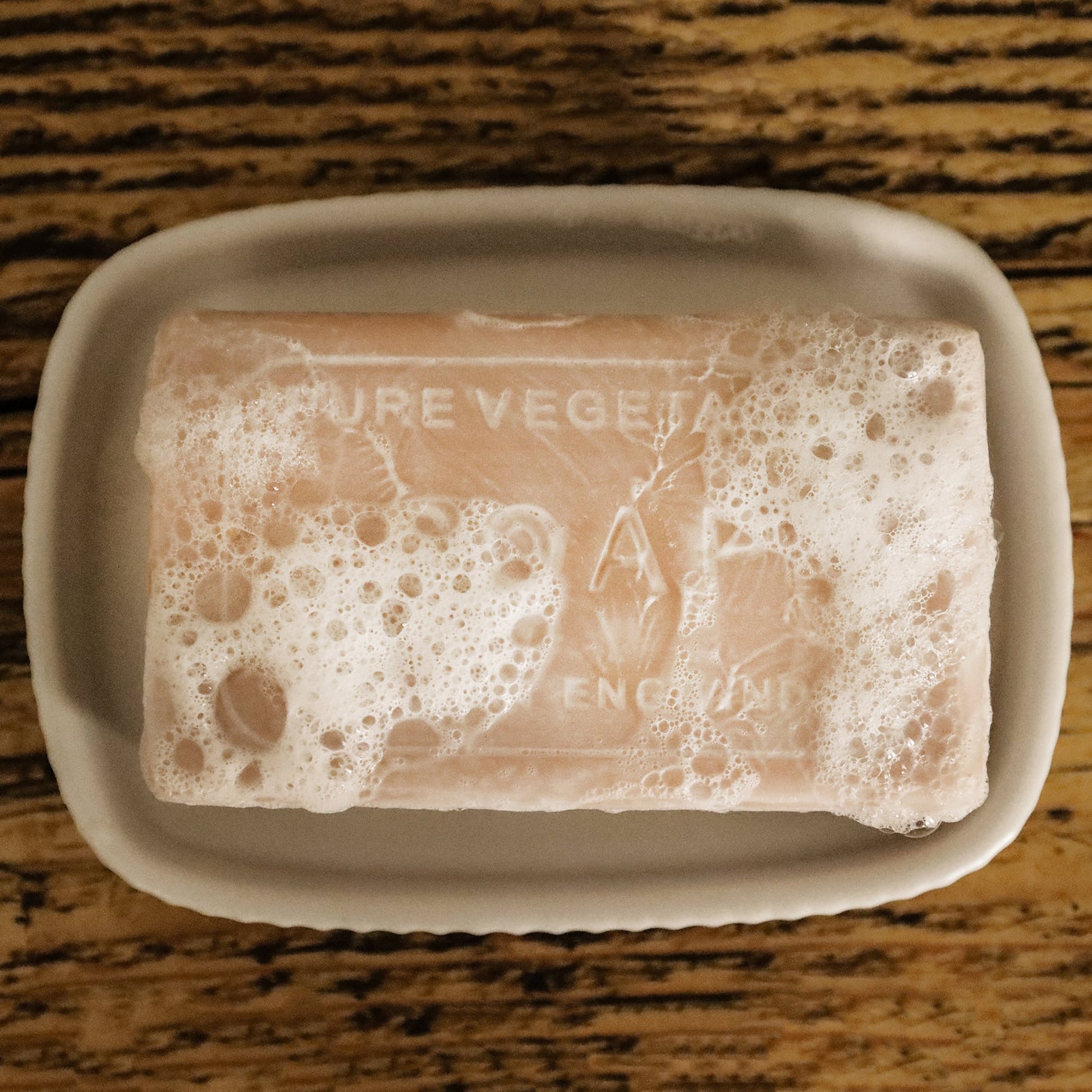 A bar of pure vegetable soap, partially covered in bubbles, sits in a simple white soap dish atop a wooden surface. The soap's engraved text 'Made in England' is visible beneath the foam.