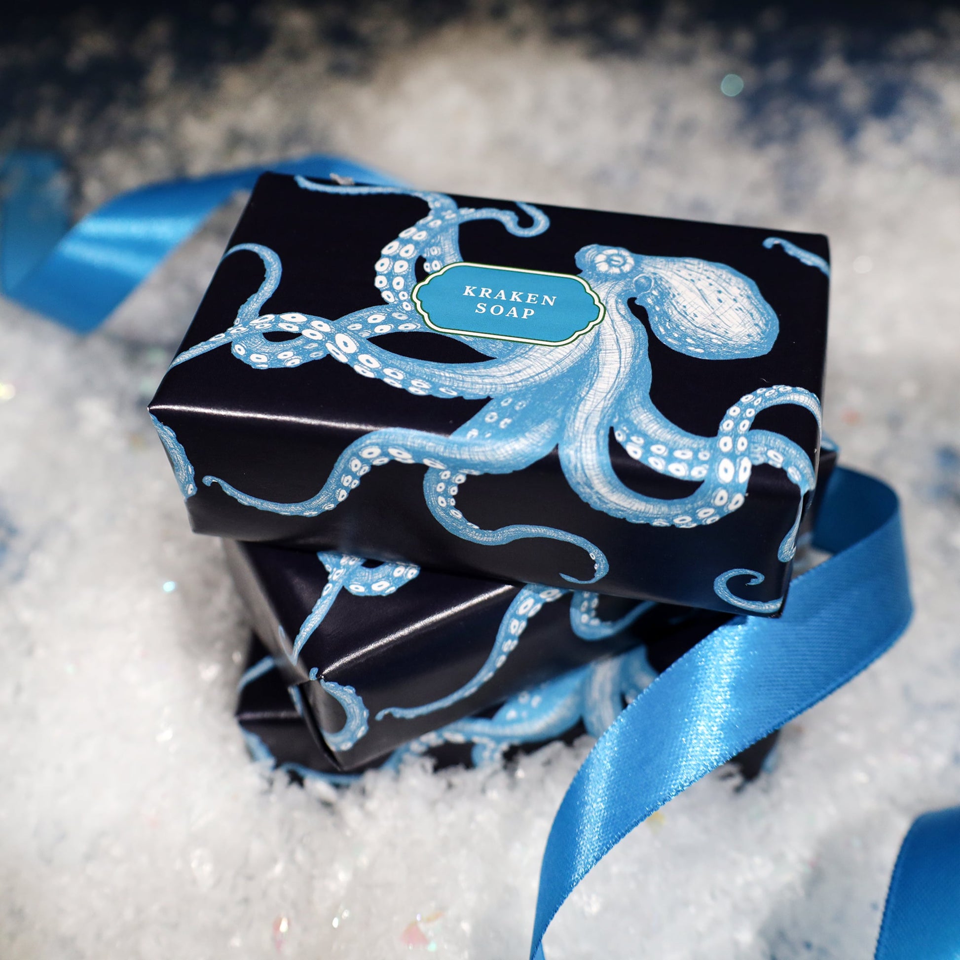 Two bars of Kraken Soap wrapped in dark navy packaging featuring a blue illustrated octopus, resting on faux snow with a satin blue ribbon curling around them in a festive winter setting.