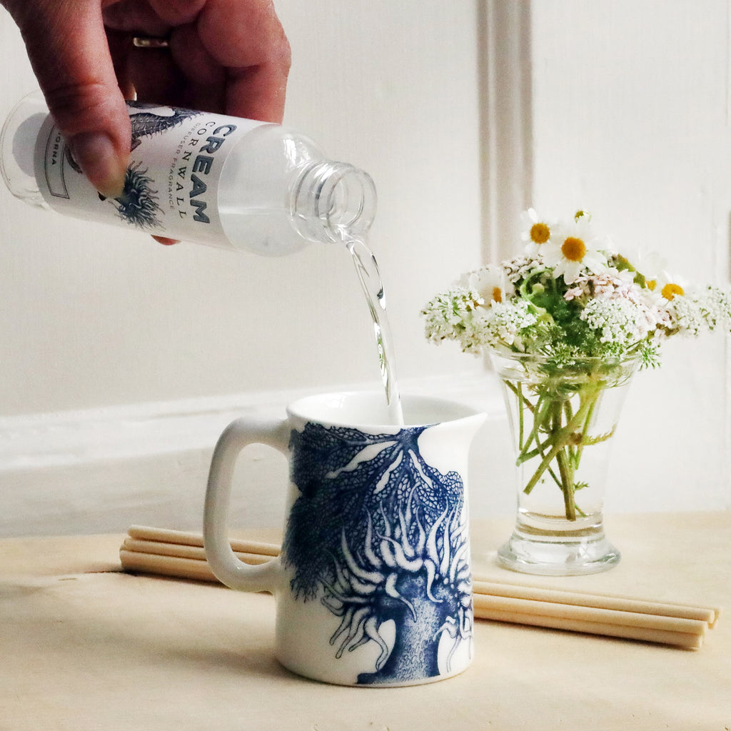 Mini Jug with sea anemone and coral design on with reeds and small posy of flowers behind with a bottle of Lamorna room diffuser oil being poured into the jug.