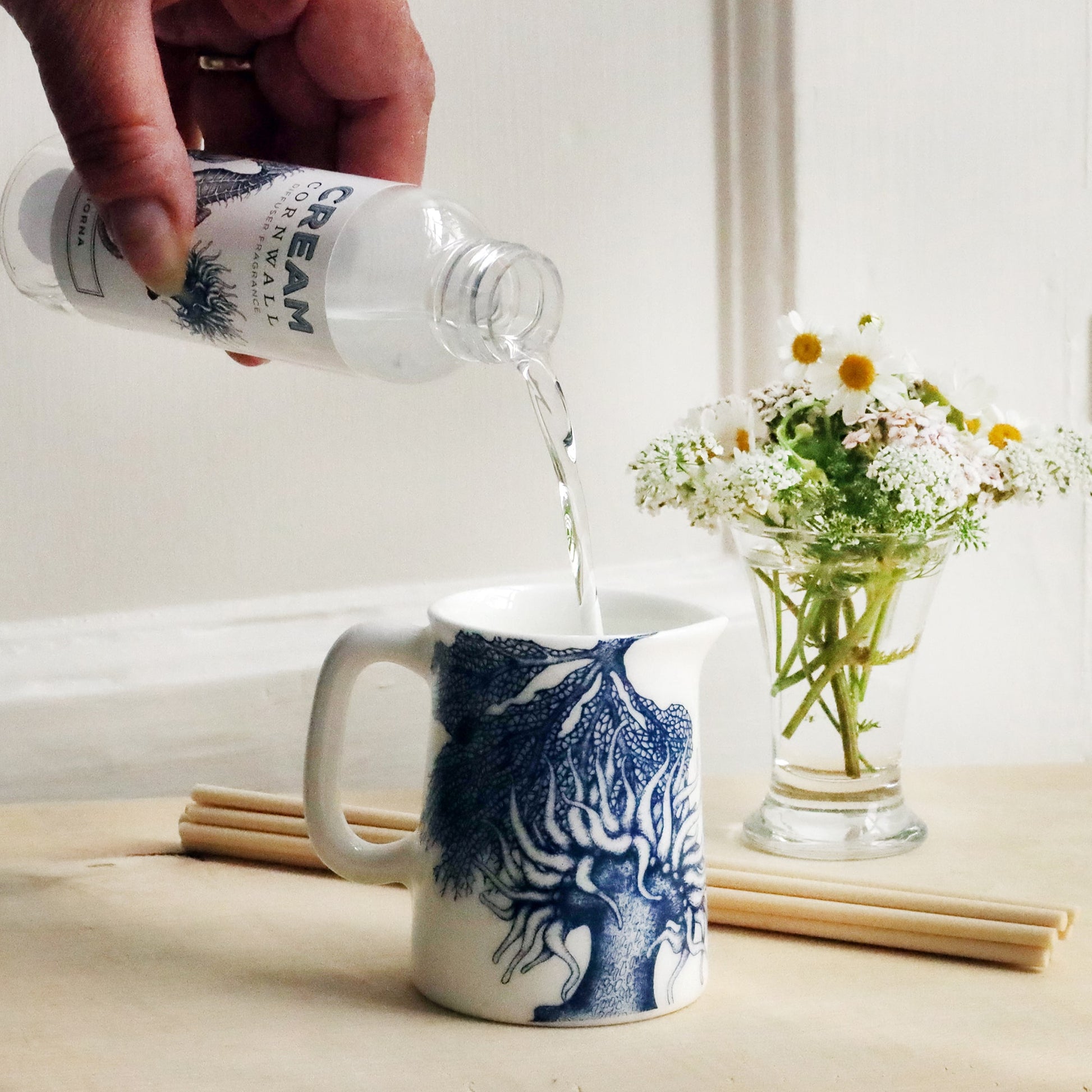 Mini Jug with sea anemone and coral design on with reeds and small posy of flowers behind with a bottle of Lamorna room diffuser oil being poured into the jug.