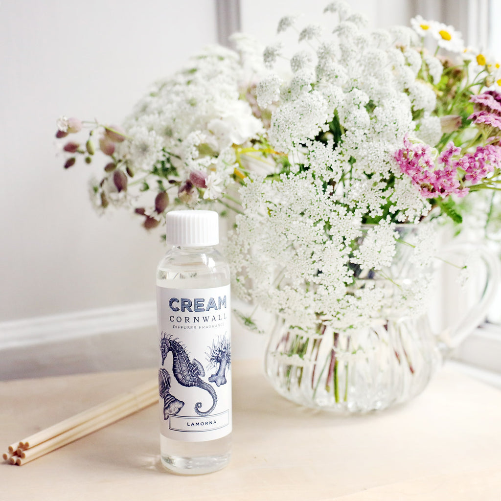 Bottle of Lamorna room diffuser oil with octopus design on the label, sitting on a wooden block with the diffuser reeds laying jut to the side, and a vase of soft coloured flowers sitting behind.