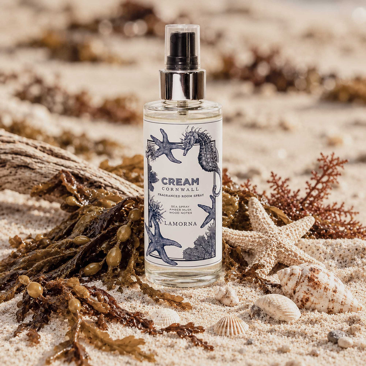 Cream Cornwall Lamorna fragranced room spray with navy seahorse and starfish illustrations, displayed on pale beach sand surrounded by seaweed, driftwood, starfish and seashells in a softly sunlit coastal setting.