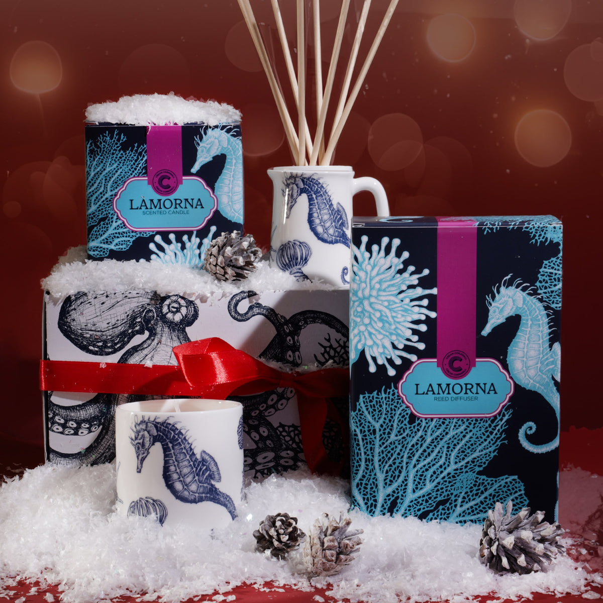Lamorna candle and reed diffuser gift hamper with seahorse and coral designs, displayed with a small ceramic jug, pinecones, and artificial snow, tied with a red ribbon against a warm festive red background with soft glowing lights.