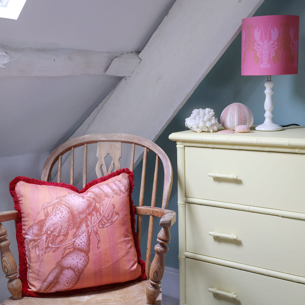 Pink pillow with a lobster design on a wooden chair next to a white dresser with a pink lamp.