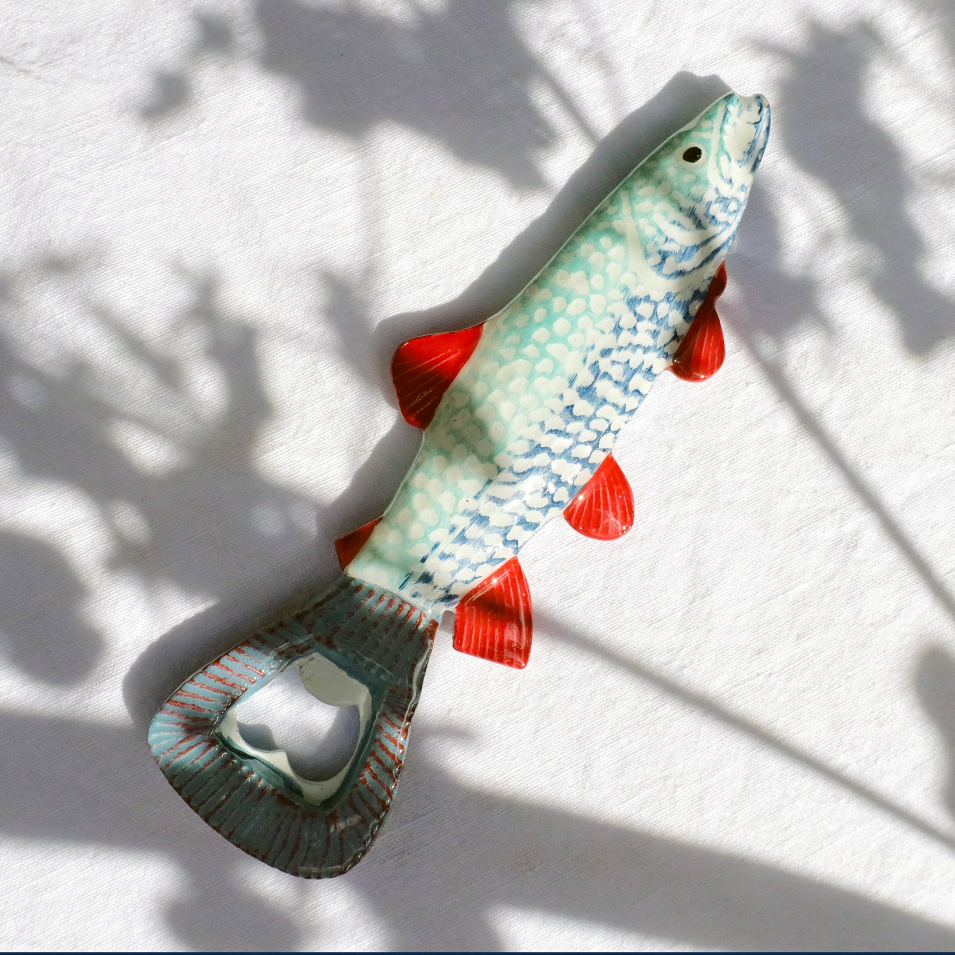 Ceramic fish-shaped bottle opener with a turquoise and blue scaled body, bright red fins, and a metal opener integrated into the tail, lying on a white fabric background with soft shadows.