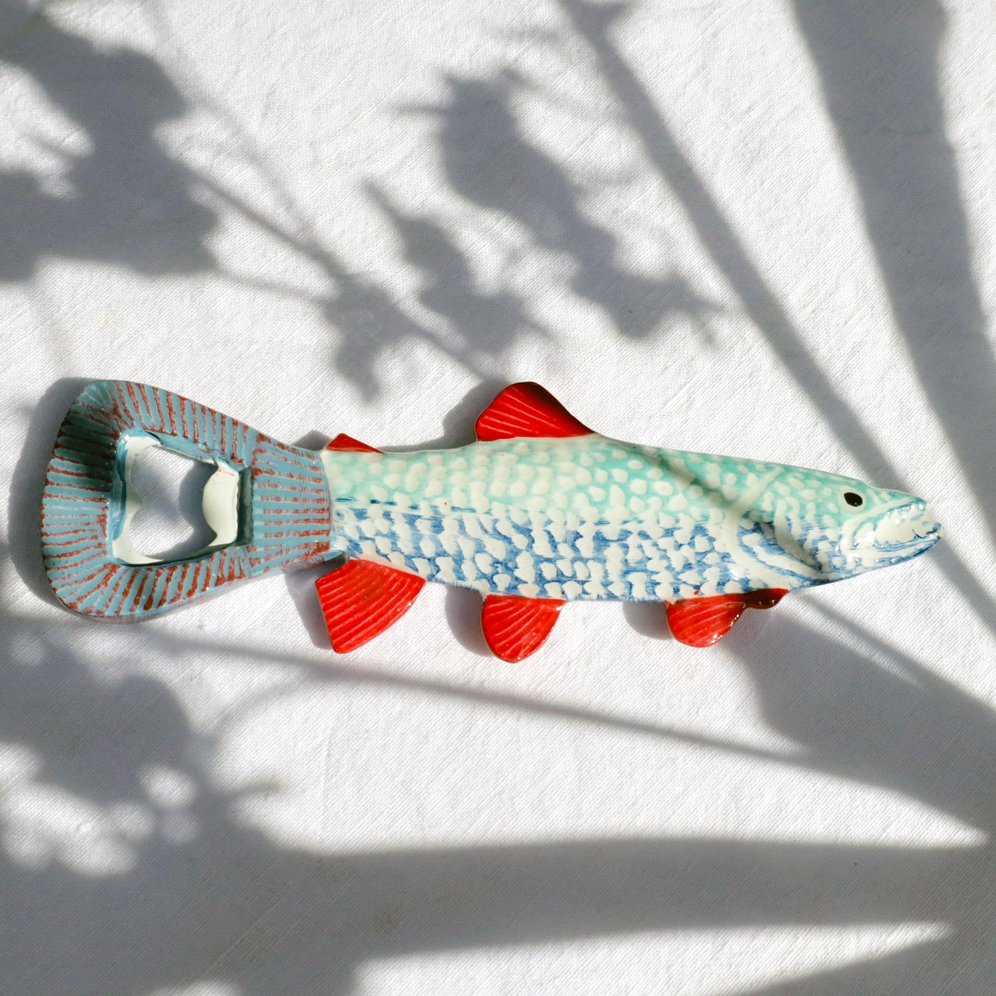 Ceramic fish-shaped bottle opener with a turquoise and blue scaled body, bright red fins, and a metal opener integrated into the tail, lying on a white fabric background with soft shadows.