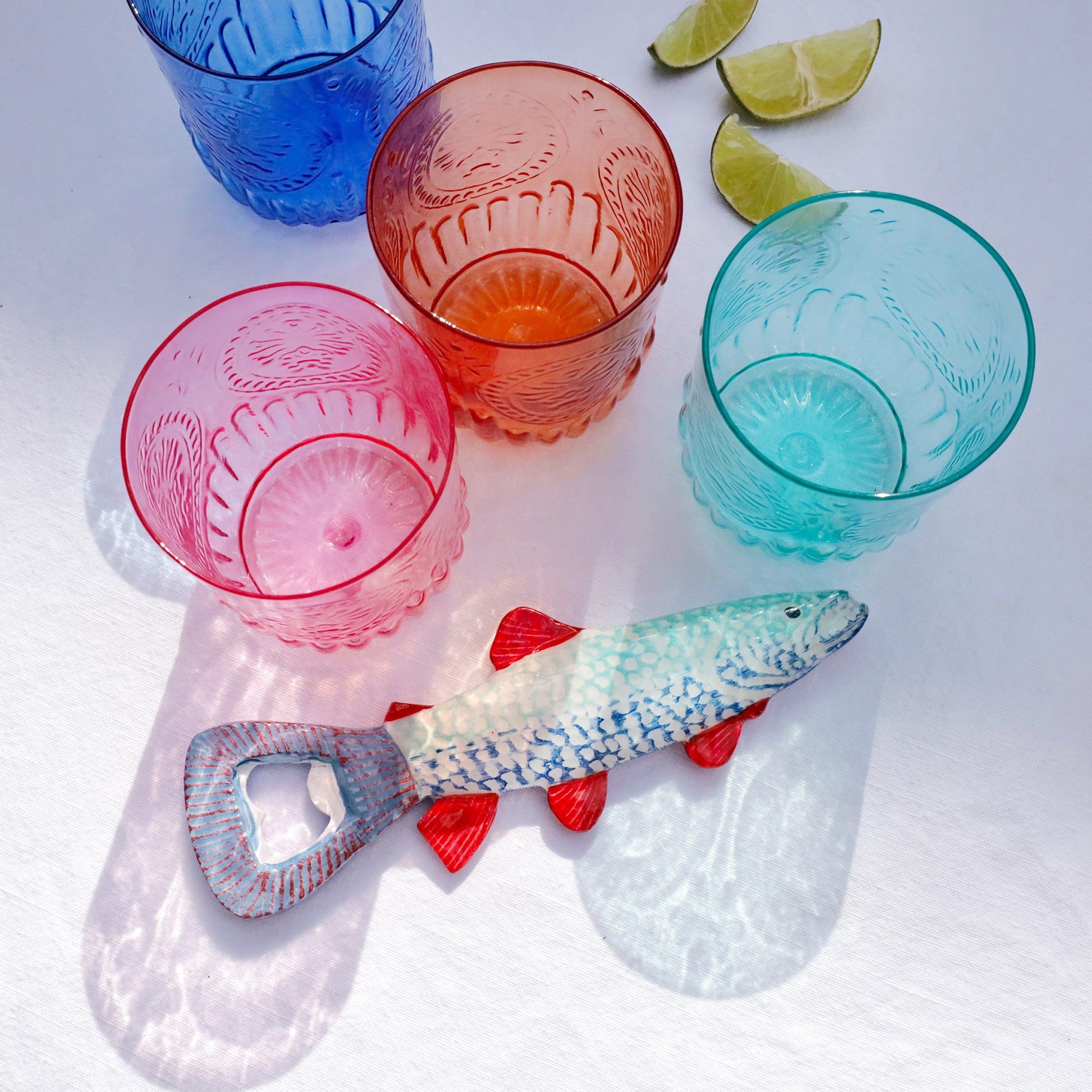Colorful fish-shaped bottle opener with blue scales and red fins placed beside four patterned glass tumblers in pink, orange, turquoise, and blue, with lime wedges in the background on a white surface.