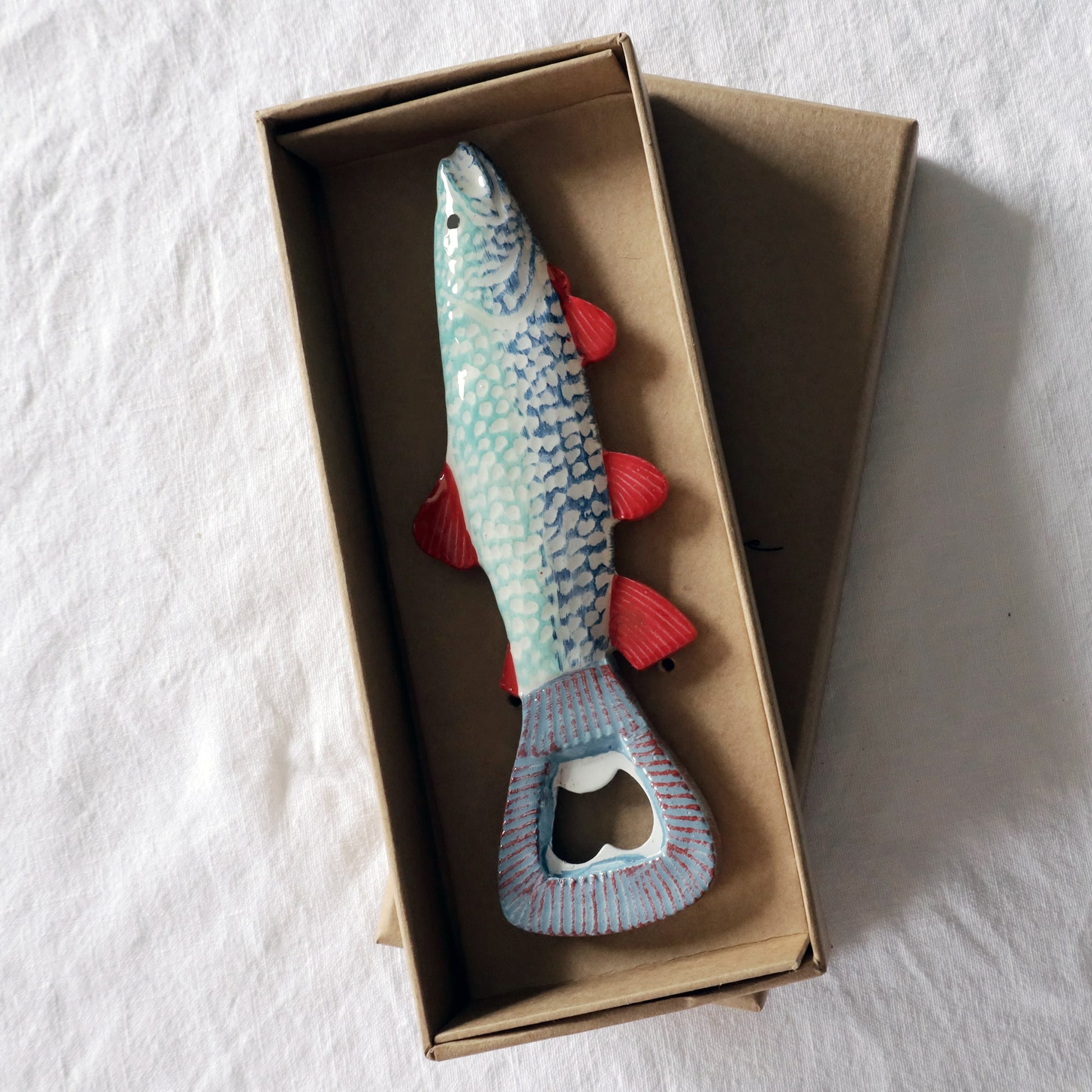 Fish-shaped bottle opener with blue scales and red fins, presented in a brown cardboard gift box on a white fabric background.