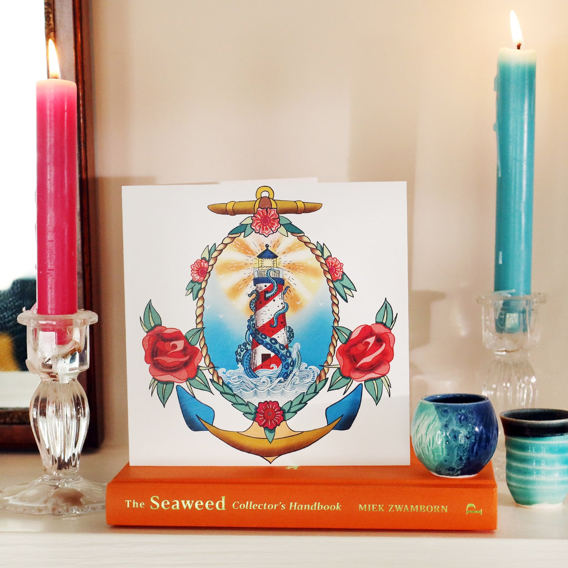 White card with illustrated lighthouse tattoo inspired illustration, sitting on a book on a shelf with a candle lit each side and some small blue ceramic pots.